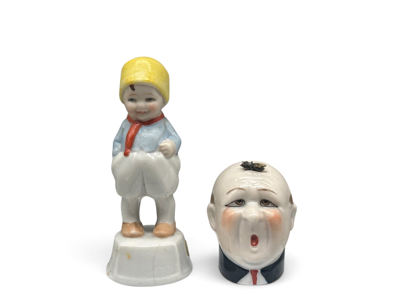 French and German Bisque Porcelain Tape Measures: Two, early 20th C. bisque porcelain tape measures to include a French figure with man's head on a fly body with cloth tape; and a German Dutch Boy figure with cloth tape. Larger: 4in tall x 1in wide. 