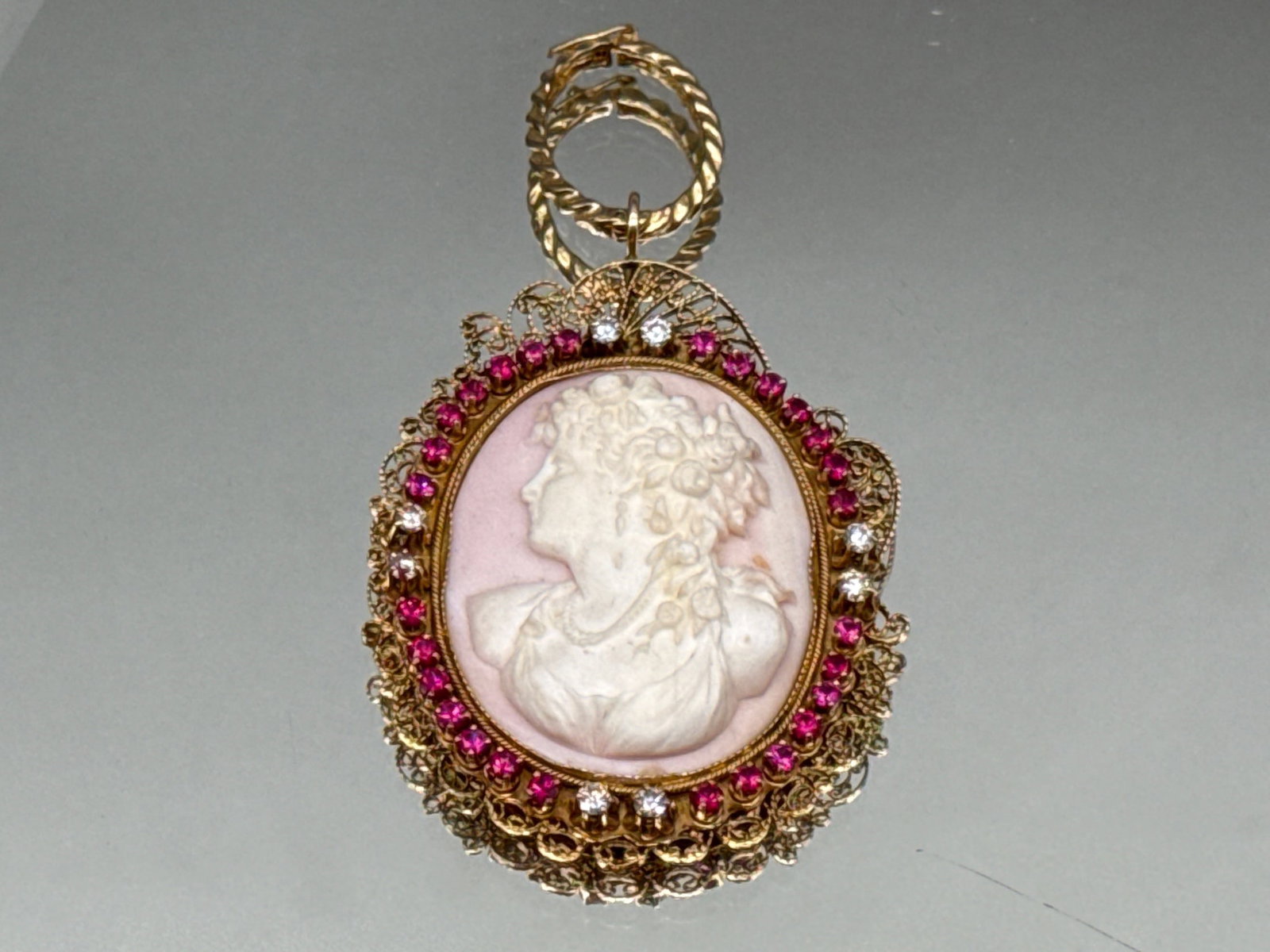 18K Diamond and Ruby Shell Carved Cameo Pendant: An 18K yellow gold filigree pendants set with a carved shell cameo with left facing neoclassical silhouette, surrounded by a halo of eight diamonds and twenty-eight rubies. Pendant: 3in tall x 2in wid