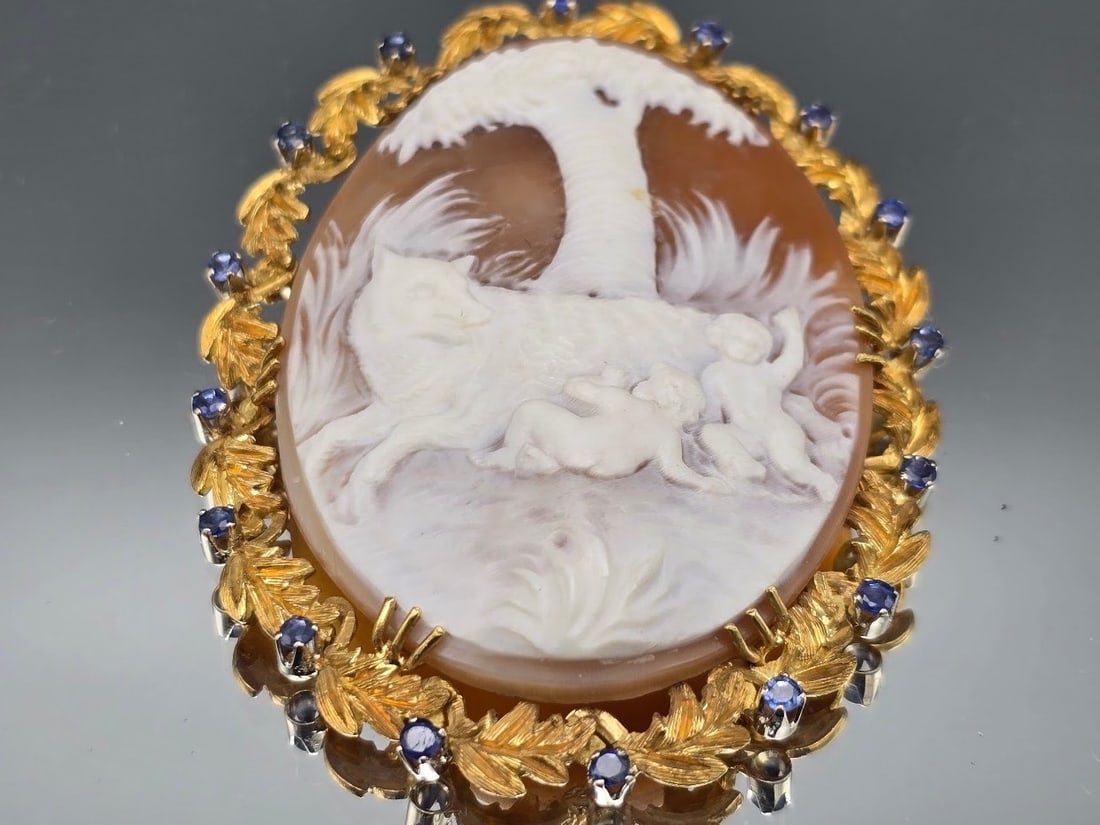 18K Carved Shell Cameo and Blue Topaz Brooch: A carved shell cameo depicting a mythological scene of cherubs and a wolf, in an 18K yellow gold setting of leaves studded with blue topaz stones. Marked Italy 18K at clasp. 3in tall x 2in wide.