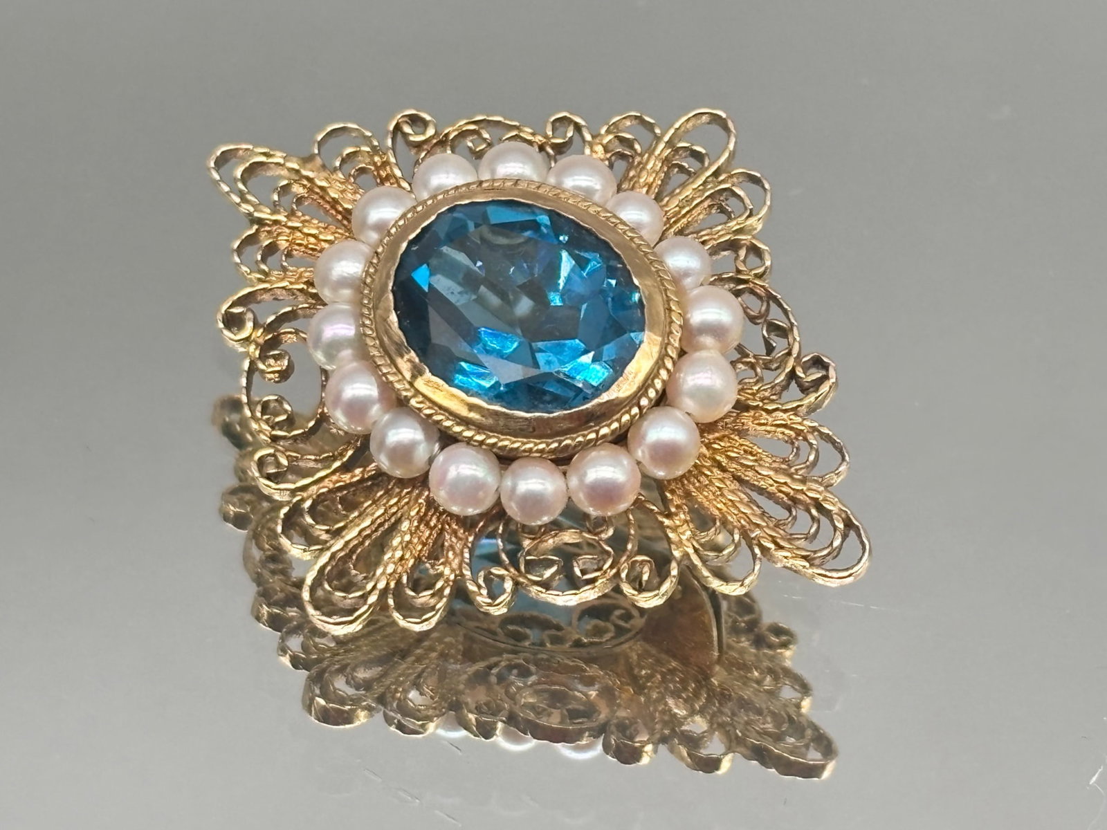 14K Blue Topaz and Pearl Brooch: A 14K yellow gold brooch of openwork filigree design with a central oval cut 5.3ct blue topaz surrounded by a halo of pearls. 1-1/2in tall x 1in wide. 5.91 dwt. | You have additional shipping