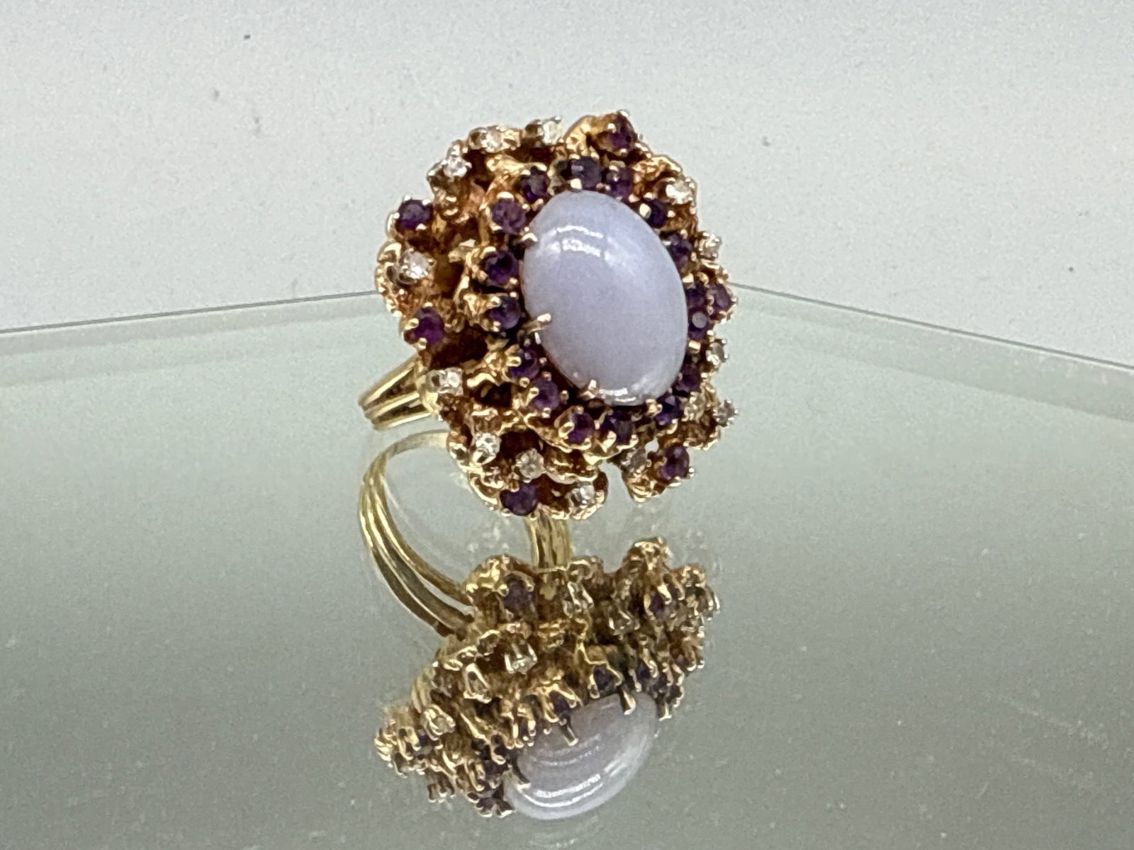 18K Purple Jade, Amethyst, and Diamond Ring: An 18K yellow gold brutalist-style cocktail ring with an oval cut purple jade stone surrounded by a halo of amethysts and thirteen diamonds. Ring size 8. 19.03 dwt. | You have additional shipping opti