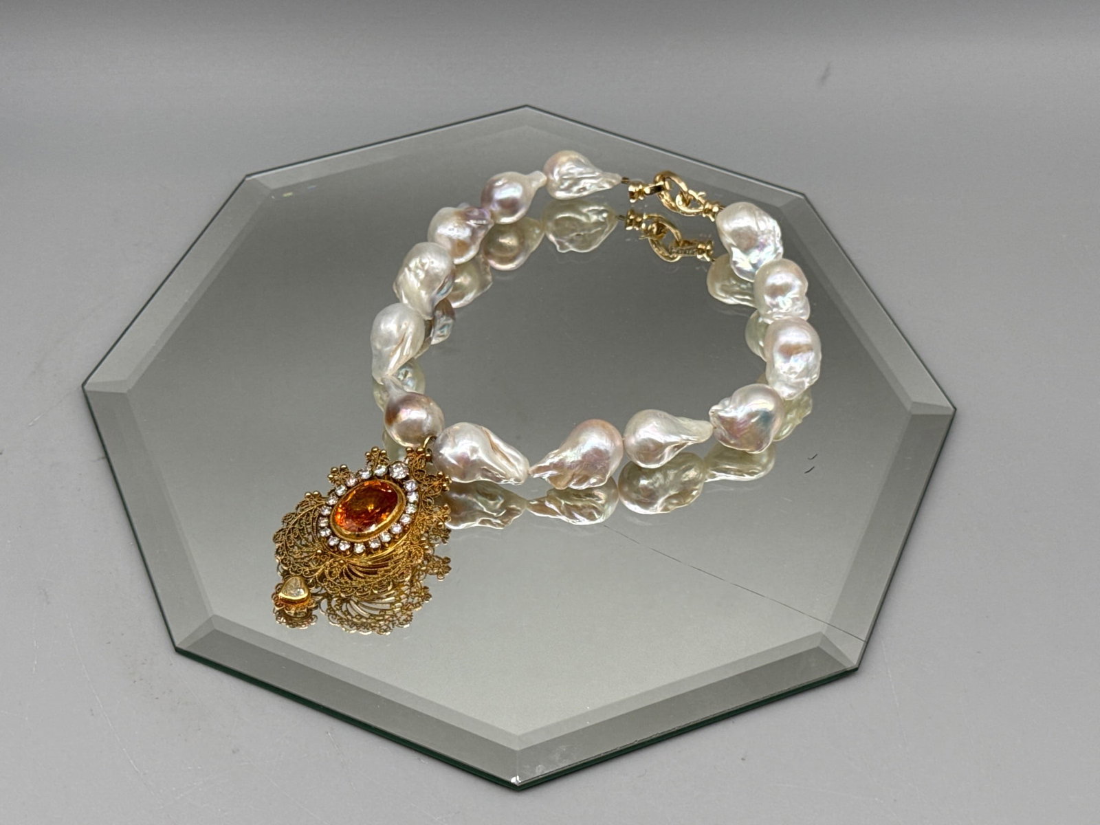 18K Baroque Pearl, Topaz and Diamond Necklace: An 18K yellow gold filigree pendant with central oval cut 12.33ct topaz surrounded by halo of graduated diamonds and with a heart-shaped diamond drop charm. Pendant is attached to a string of