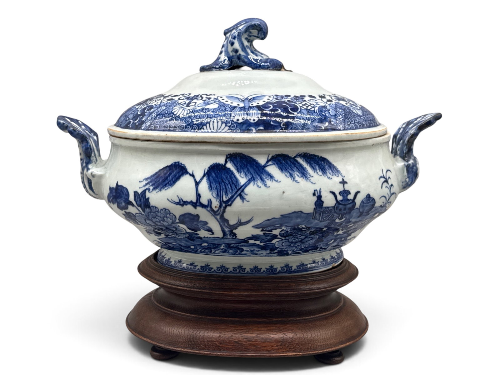 Chinese Export Blue & White Lidded Tureen: A Chinese export porcelain blue and white covered soup tureen with botanical scenes, butterfly motifs, and leaf-form handles resting on a wooden plinth. 10in tall x 12in wide. | You have additional sh