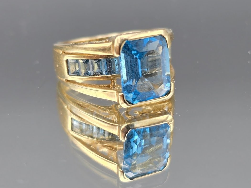 14K Ring with Emerald Cut Blue Topaz: A 14K yellow gold cocktail ring with raised filigree shoulders mounted with baguette cut blue topaz accents surrounding a central emerald cut blue topaz. Marked 14K. Size 7. 3.34 dwt. | You have