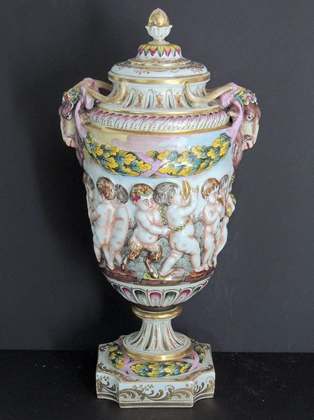Capodimonte Urn: Capodimonte polychrome-decorated three-part urn with lid. Featuring two ram’s head handles and embossed putti musician design on body. Marked with blue “N” with crown mark. 15-1/2” tall, 8”
