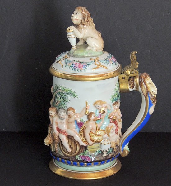 Capodimonte Tankard: Capodimonte polychrome-decorated tankard, featuring embossed classical figures and animals, lion finial, and goat handle. Brass and porcelain lid; gilt interior. Marked with blue “N” with crown ma