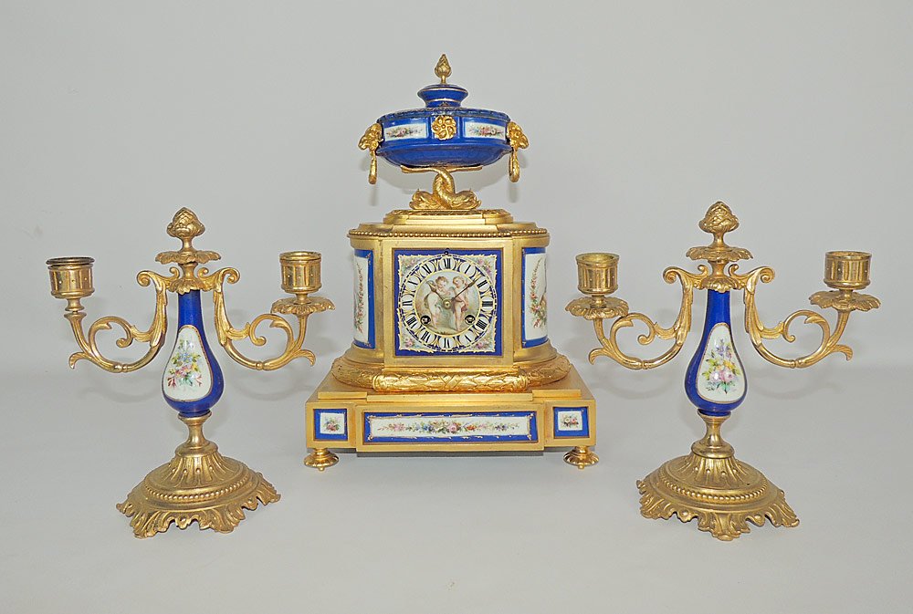 French Bronze and Porcelain Clock Garniture Set: French gilded-bronze and porcelain mantel clock garniture set. Clock features a gilded-bronze base with 8 porcelain putti, floral, and portrait-decorated panels, with a ram's head porcelain finial sup