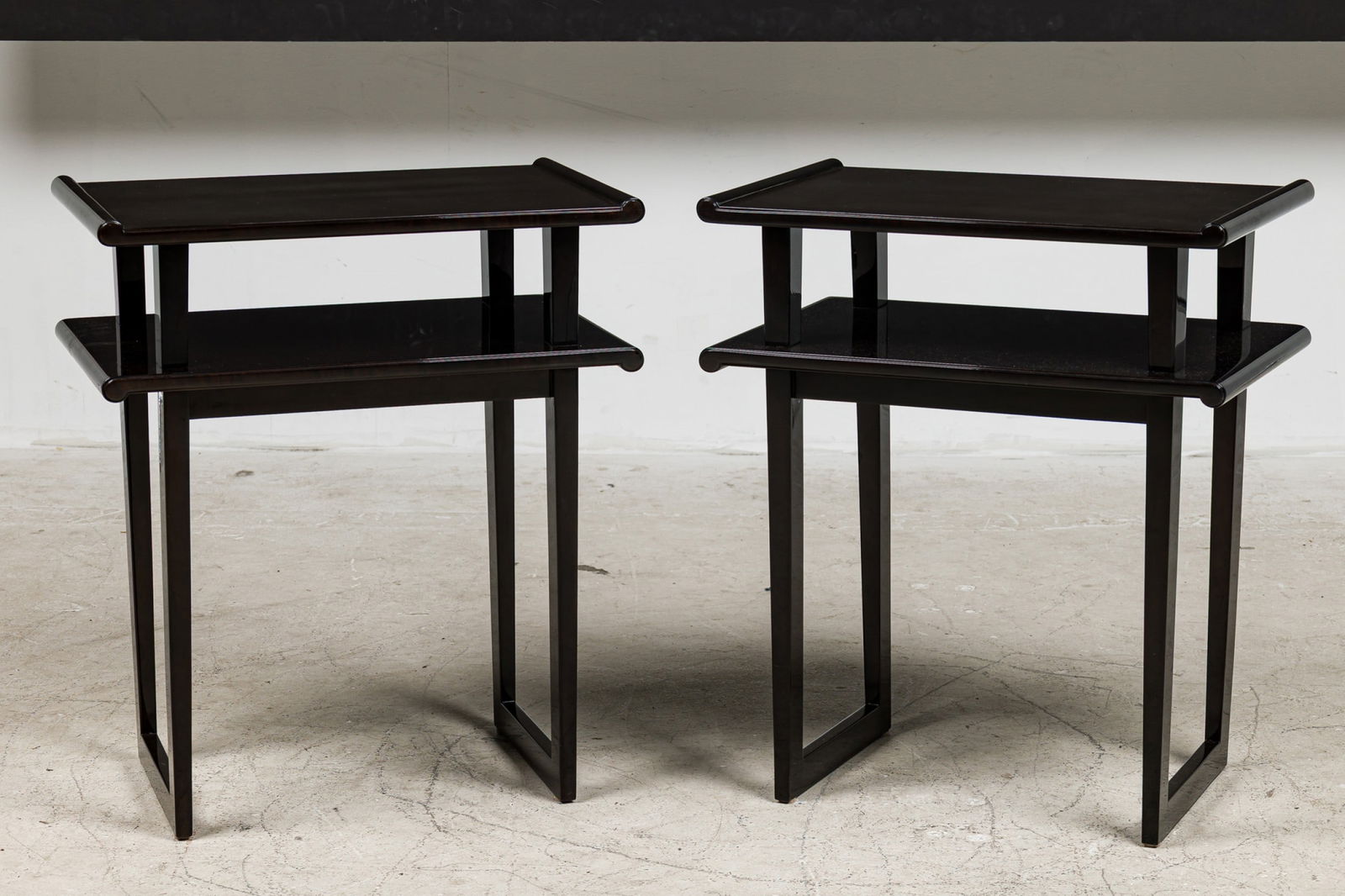 James Mont (Attr), Pair Art Deco Tiered Side Tables: 20th c., dark stained wood in high gloss clear lacquer, the rectangular tops with scrolling edge, raised on open trestle legs, sourced by Bunny WIlliams Interiors, scanned invoice attributes to James