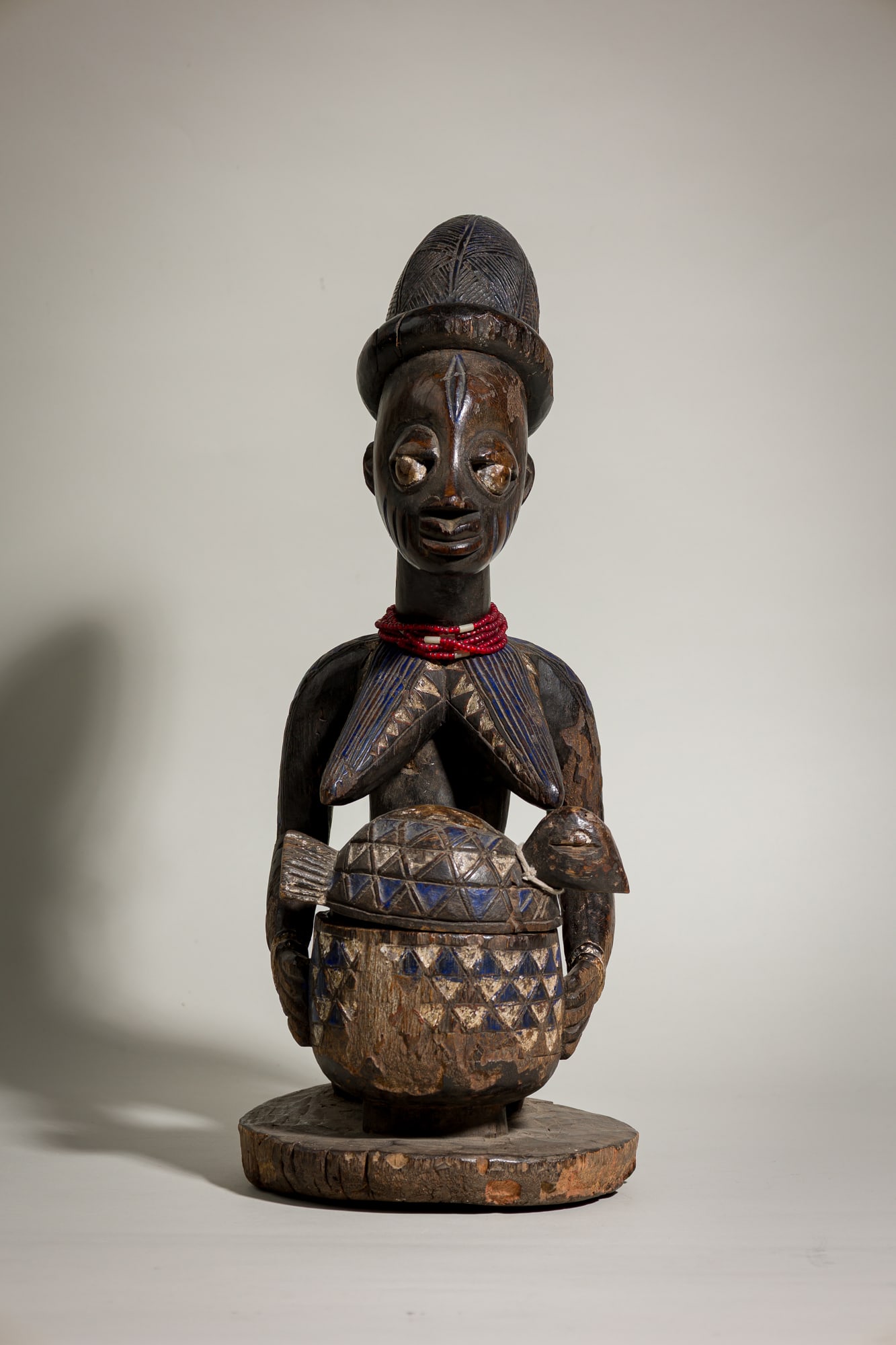 Very Old Yoruba Female Bowl Bearer Olumeye: early 19th c., Nigeria, carved wood, modeled askneeling female holding a bowl, with scarified markings on face and bust, white and blue pigments, beaded necklace, and a bird-shaped lid with geometric