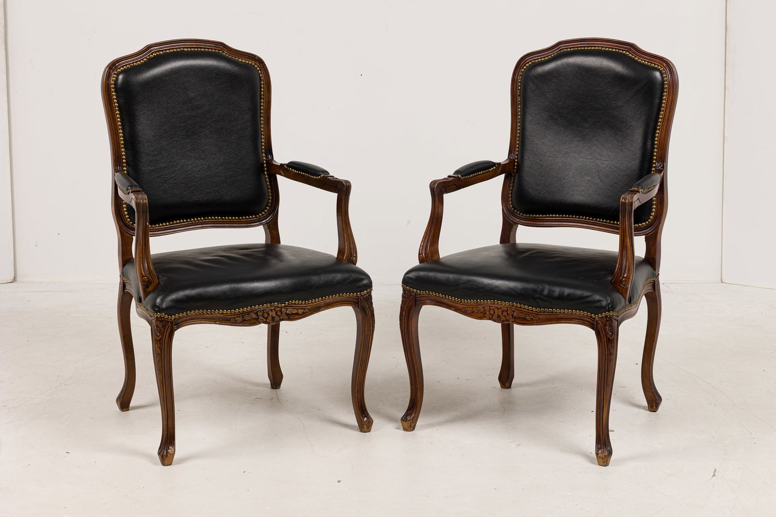Pair French Provincial style black leather armchairs: 20th c., USA, carved oak frames, seat, back, and armrests upholstered in black leather with nailhead trim, carved apron with floral design, rests on four fluted legs, 41.5"h x 24"w x 20.75"d, 18"h (to