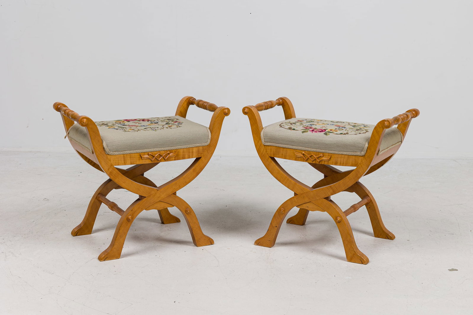 Pair French Empire style curule stools: 19th/ 20th c., France, birch wood X-shaped legs with a central carved geometric motif along the rail, needlepoint tapestry upholstered seats with floral wreath design, placed by Stephen Bast