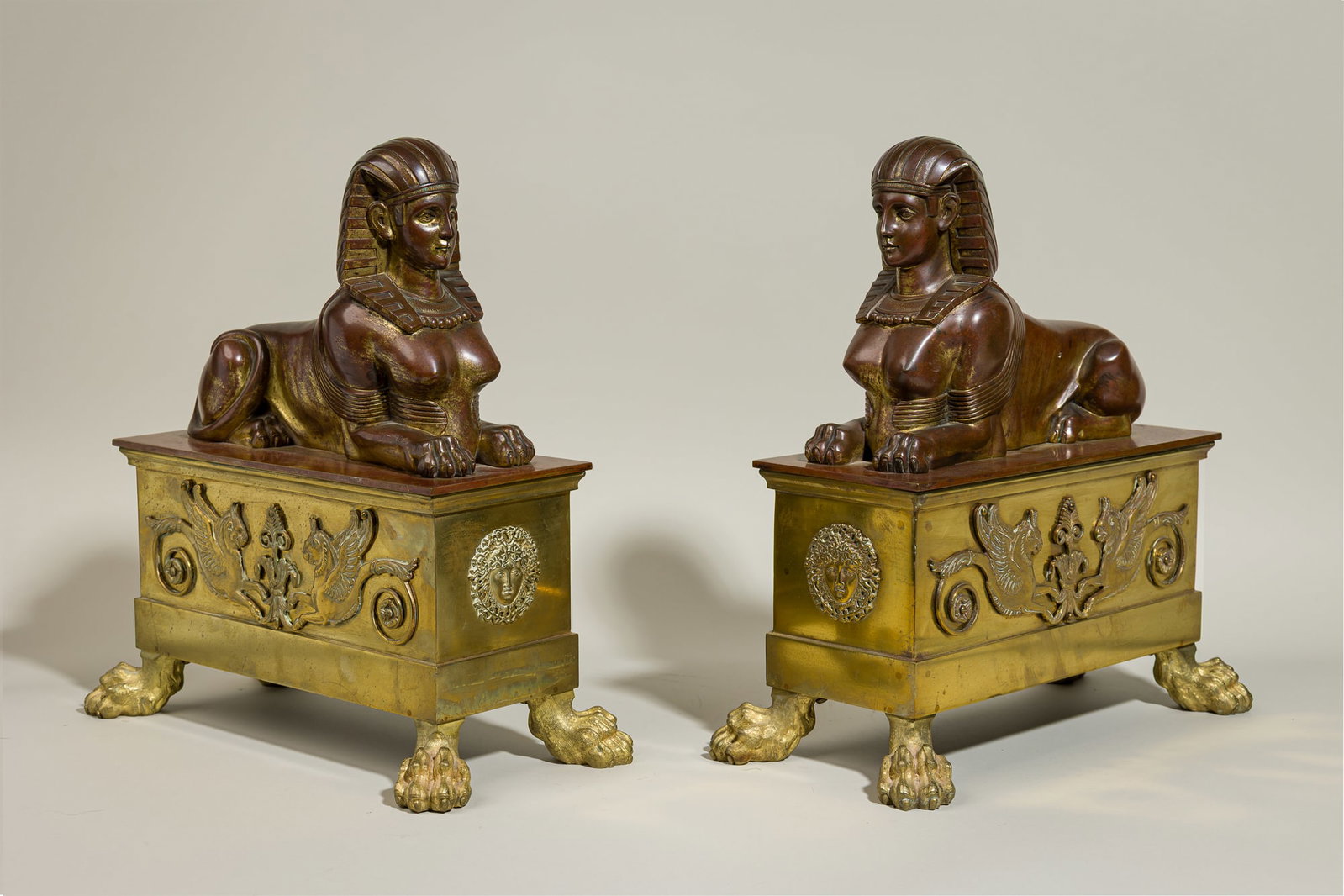Pair French Empire parcel gilt bronze recumbent sphinx chenets: 19th c., France, reddish brown patinated bronze, residual gilt finish, the gilt brass pedestals decorated with relief masks and griffins, 15"h x 7.25"w x 14"d Provenance: Pro