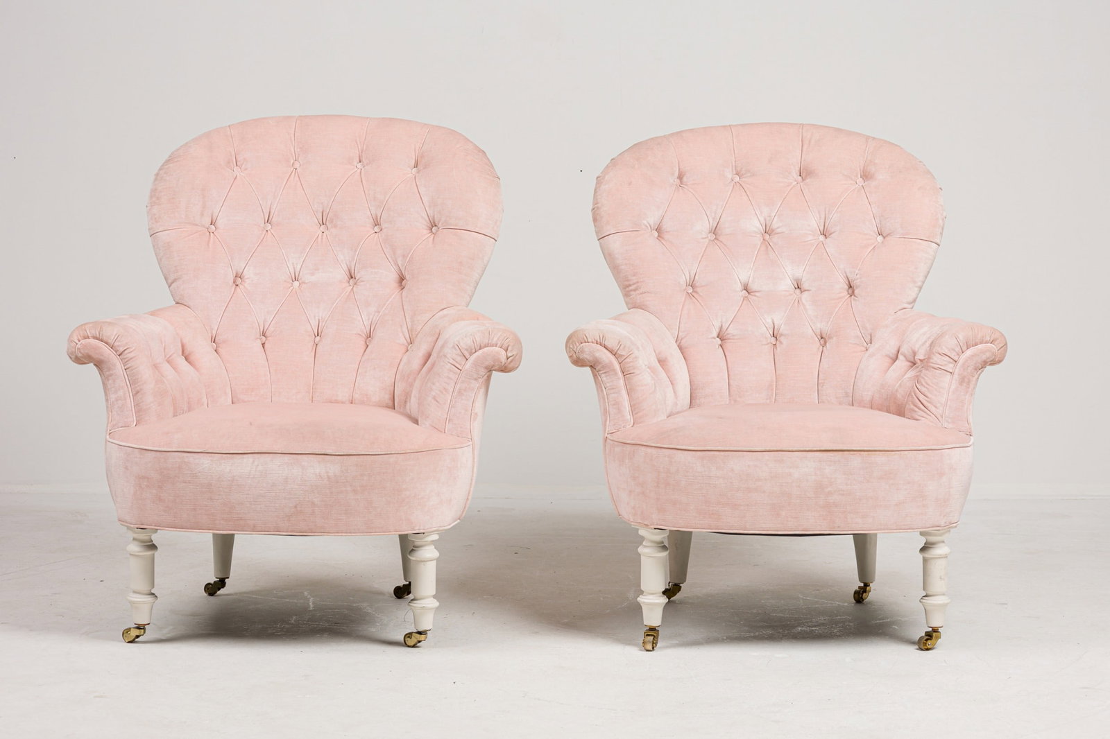 Stephen Bastone, custom capitonne armchairs: 21st c., USA, button tufted back and outward sweeping arms, upholstered in blush pink velvet, on white painted wood turned legs and brass casters, designed by Stephen Bastone