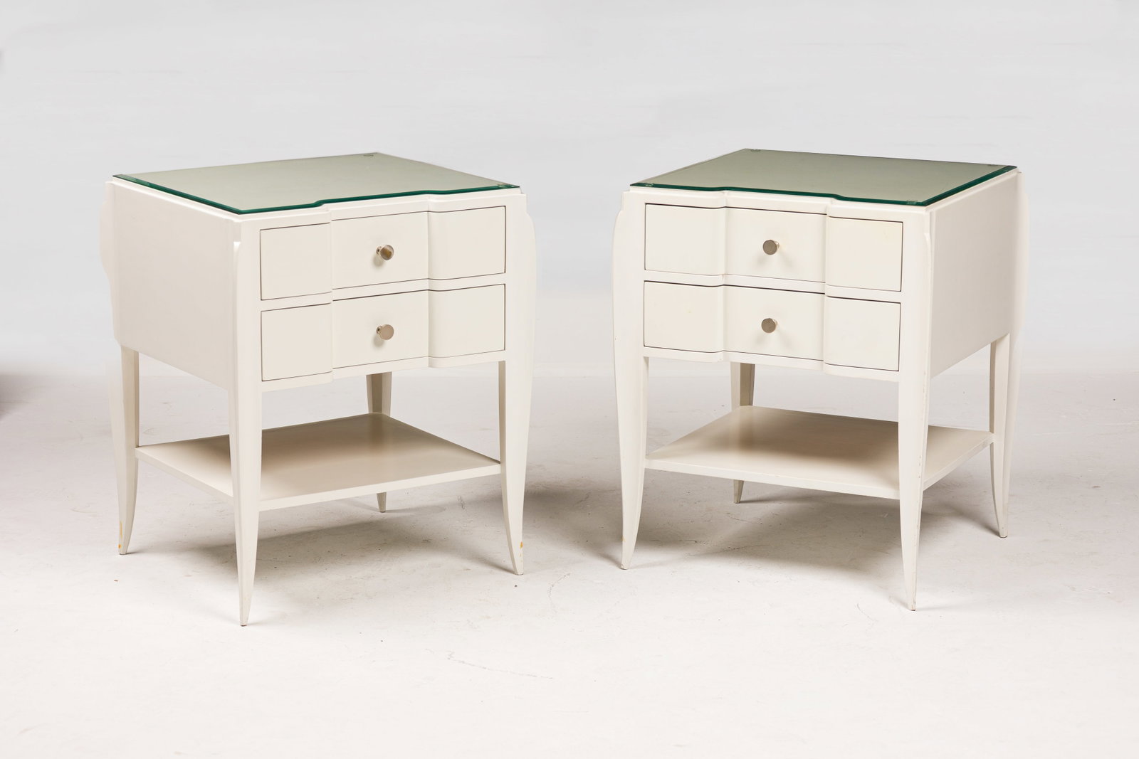 Stephen Bastone, pair, custom Art Deco style bedside tables: 21st c., USA, white lacquered wood frame, with inset glass top over two stacked drawers, the sabered legs joined by lower open shelf, mounted with brushed chrome pulls and sabots, designed&n