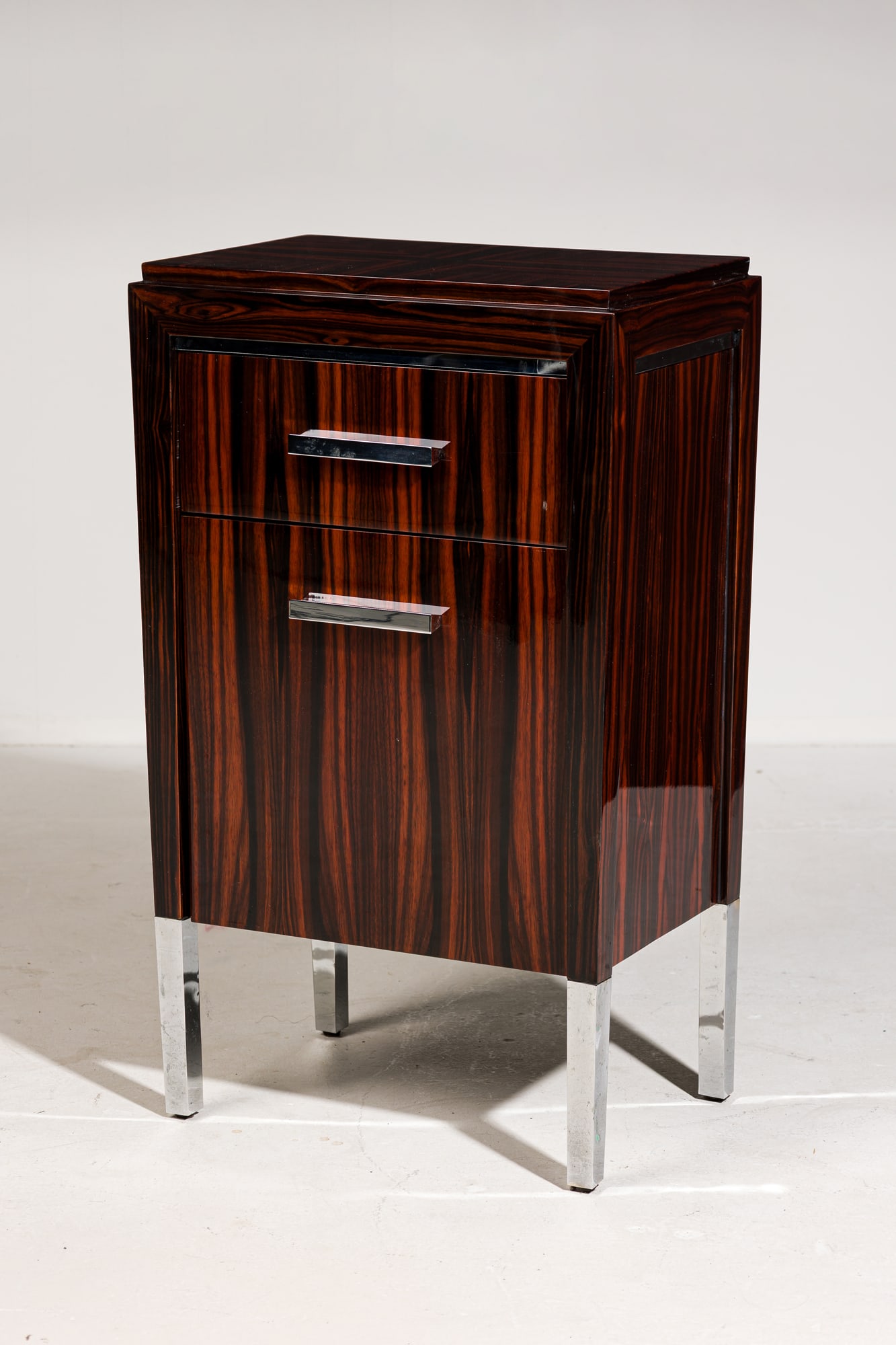 French Art Deco style Macassar Ebony small cabinet, Stephen Bastone: 21st c., USA, high polished clear lacquered Ebony veneer, the stepped rectangular top over frieze drawer and lower door opening to shelf storage, fitted with chromed metal pulls and square tube metal&