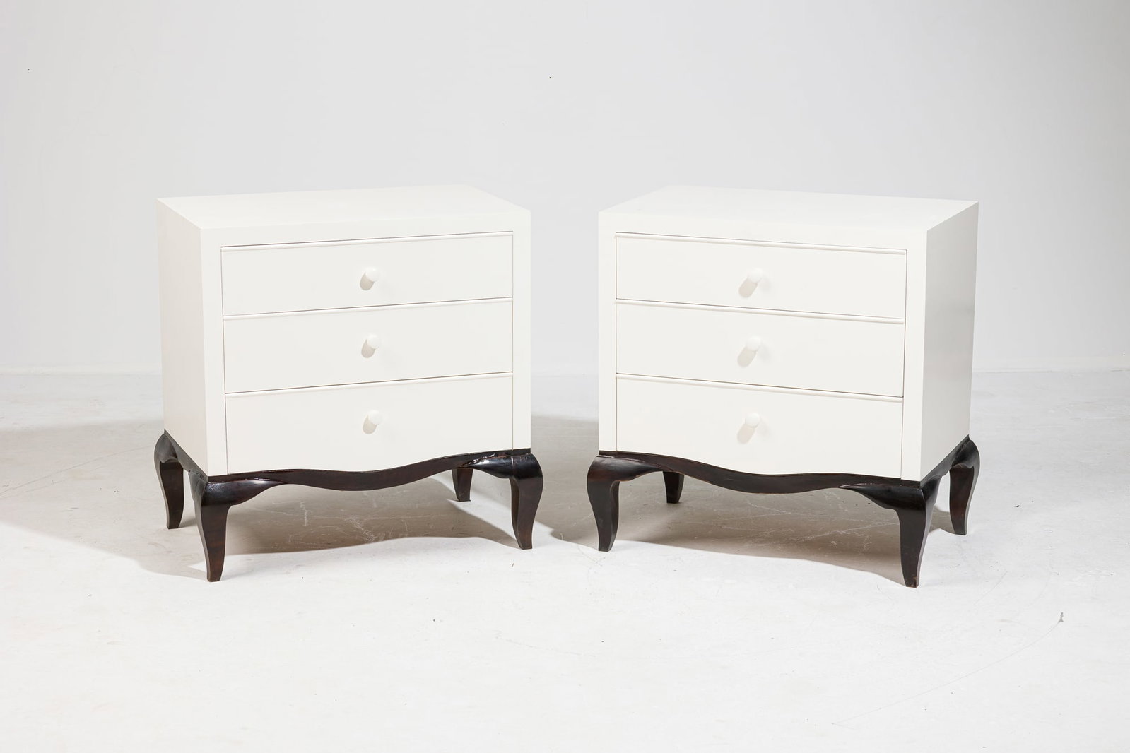Pair custom Art Deco style white lacquer nightstands, Stephen Bastone: 21st c., USA, dark stained and white painted wood, the cabinet fitted with three drawers set with wooden knobs, raised stylized cabriole legs, 28" h x 26.5" w x 16.5" d&n