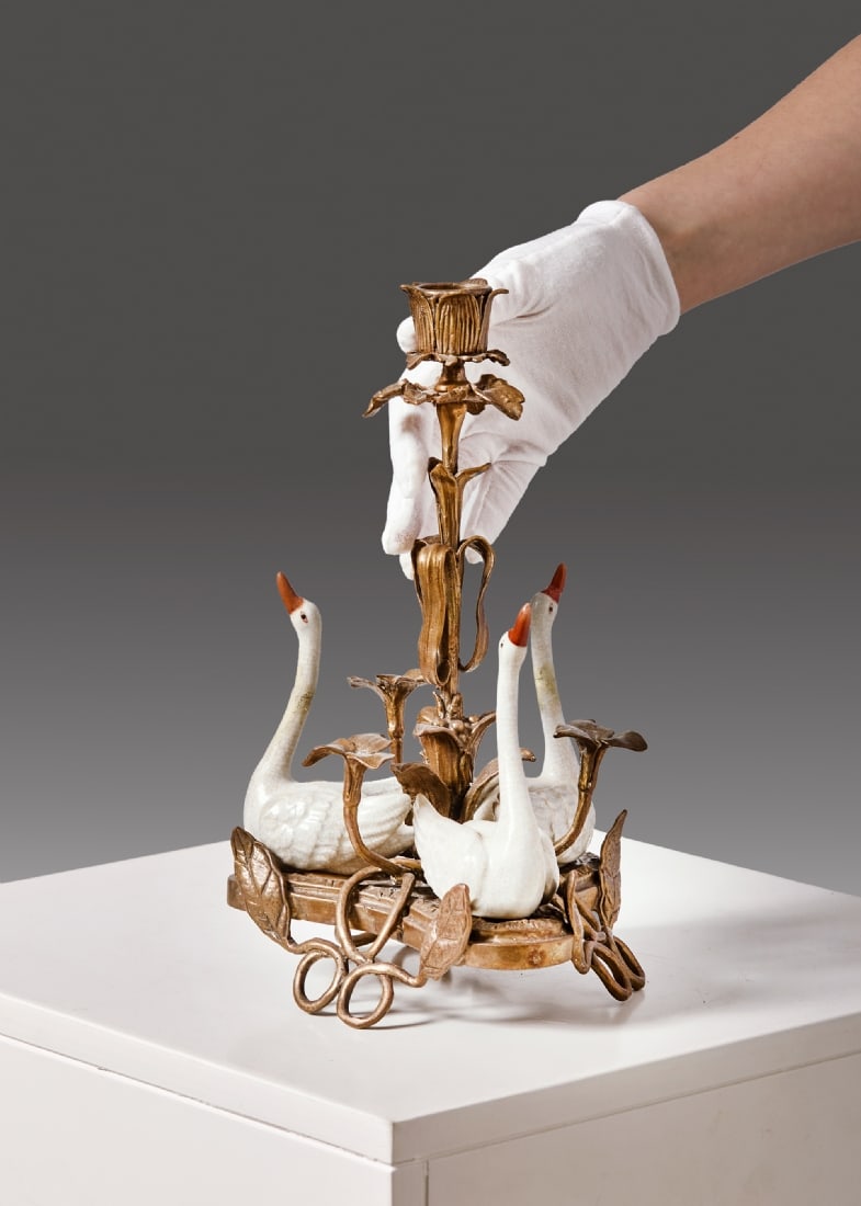 Bronze Candlestick Surrounded by Three Porcelain Swans: Bronze Candlestick Surrounded by Three Porcelain Swans, in the center stands a column adorned with leaves and flowers, holding a single candle holder Overall height: 30 cm Width: 18 cm Repair to 3 nec