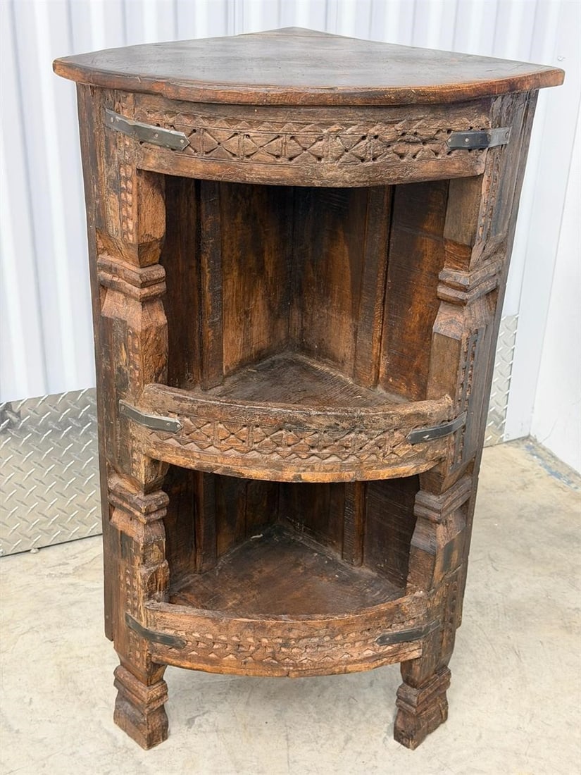 Rustic Indian Corner Display Cabinet: Rustic Indian corner display cabinet featuring carved geometric motifs and a weathered wood construction. Features: Bow front shelves, carved decorative patterns, and dark metal hardware bracing.Purpo