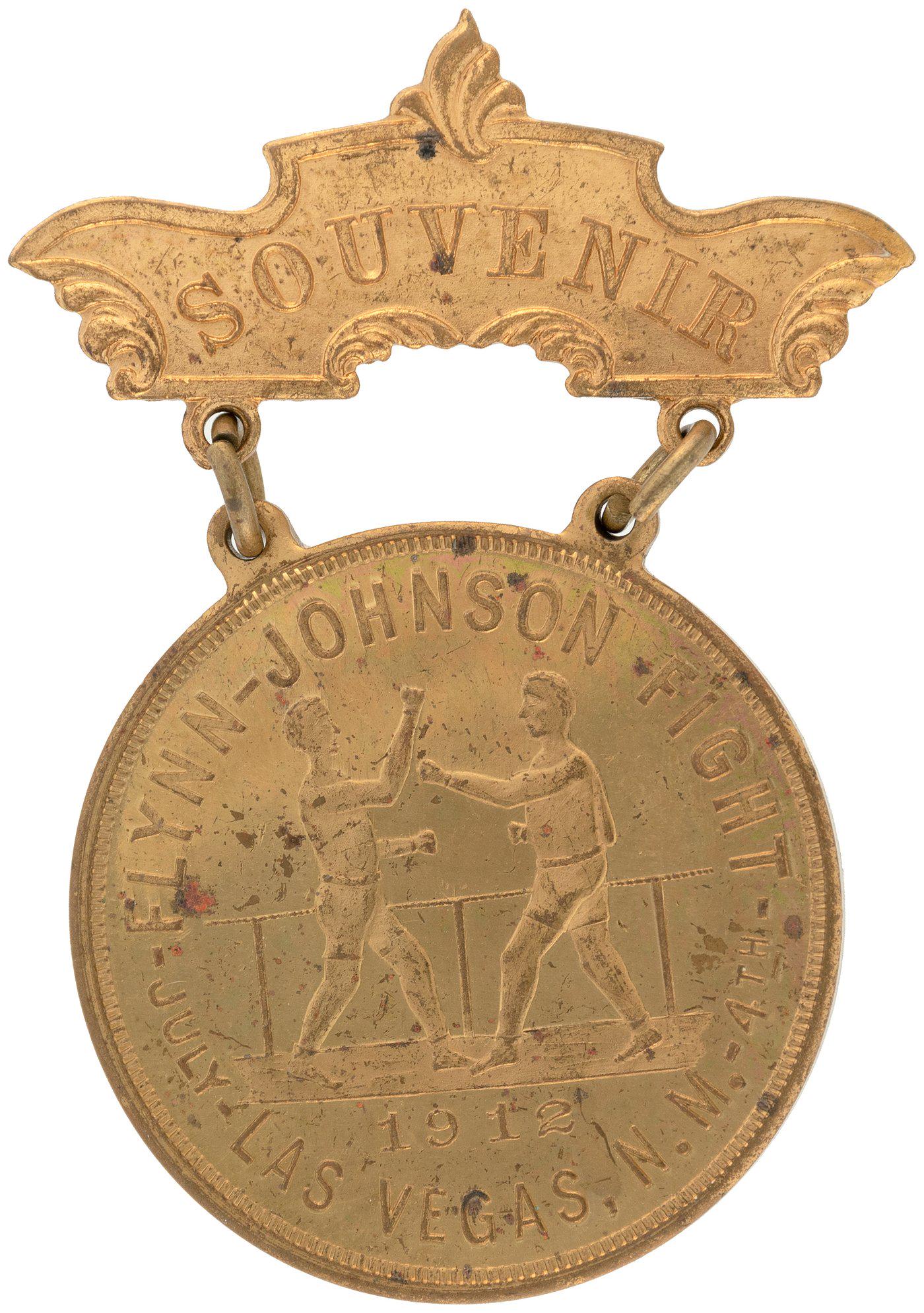 1912 BRASS BADGE FOR LAS VEGAS, NM HEAVYWEIGHT MATCH: 1912 BRASS BADGE FOR LAS VEGAS, NM HEAVYWEIGHT MATCH BETWEEN JIM FLYNN AND JACK JOHNSON. 2-5/16" tall all brass with close-up showing American flag on reverse. Obverse has fighters in a ring with "191