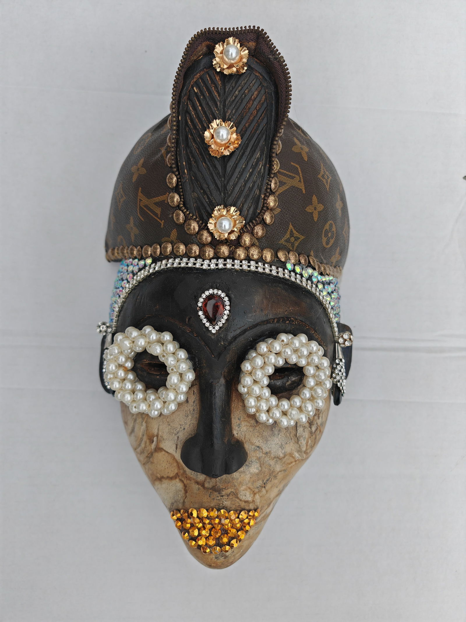 African Antique Mask Reimagined by Mbeng Pouka created in 2024 Wood and Mixed Media Artwork Not: This unique mask sculpture merges traditional craftsmanship with modern luxury, creating a striking interplay of textures and materials. Adorned with pearls, gemstones, and metallic accents, the piece
