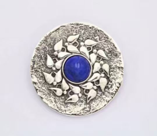 DECORATIVE JEWELRY BROOCH/PENDANT: DECORATIVE JEWELRY BROOCH/PENDANT Description: Circular decorative object in a silver-toned metal, centered by a vivid round blue cabochon surrounded by a wreath-like cluster of raised leaf and