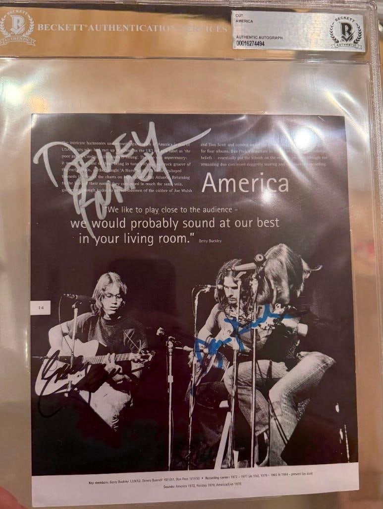 America Fully Group Signed Slabbed 8" x 6" Book Photo Cut (3 Sigs) (Beckett/BAS Authentication): Black-and-white slabbed 8" x 6" cut signed on the front in silver, black, and blue felt tip markers by America band members Dan Peek, Dewey Bunnell, and Gerry Buckley. In fine condition. Encapsulated