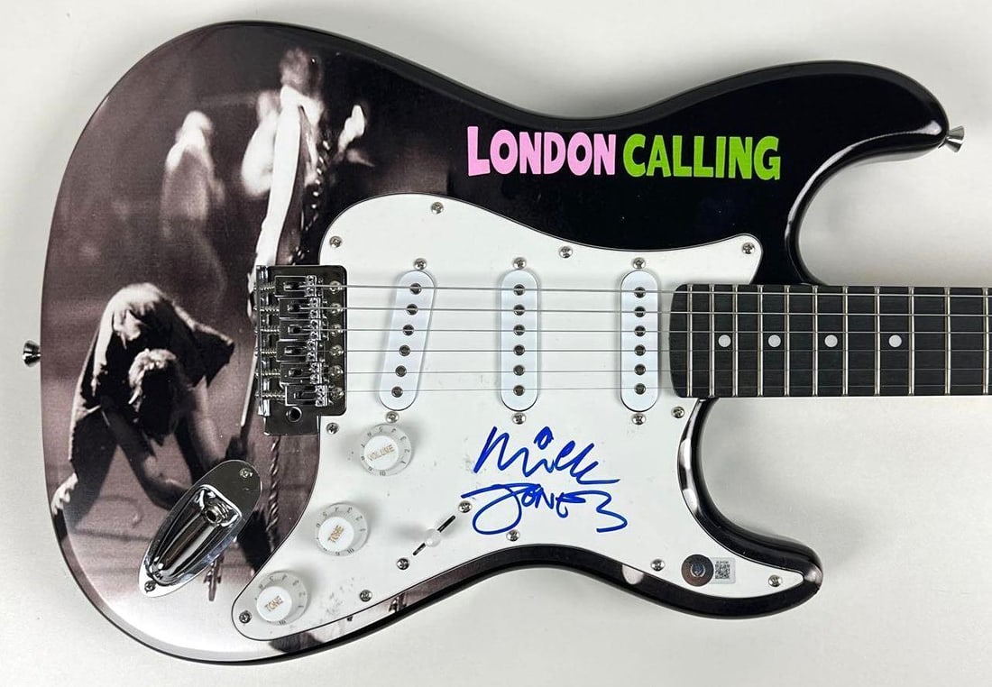 The Clash: Mick Jones Signed "London Calling" Custom Stratocaster-Style Guitar (Beckett): "London Calling" custom graphics Stratocaster-style guitar signed on the pickguard in blue felt tip marker by The Clash band member Mick Jones. Measuring approximately 39" x 13" and in fine condition.