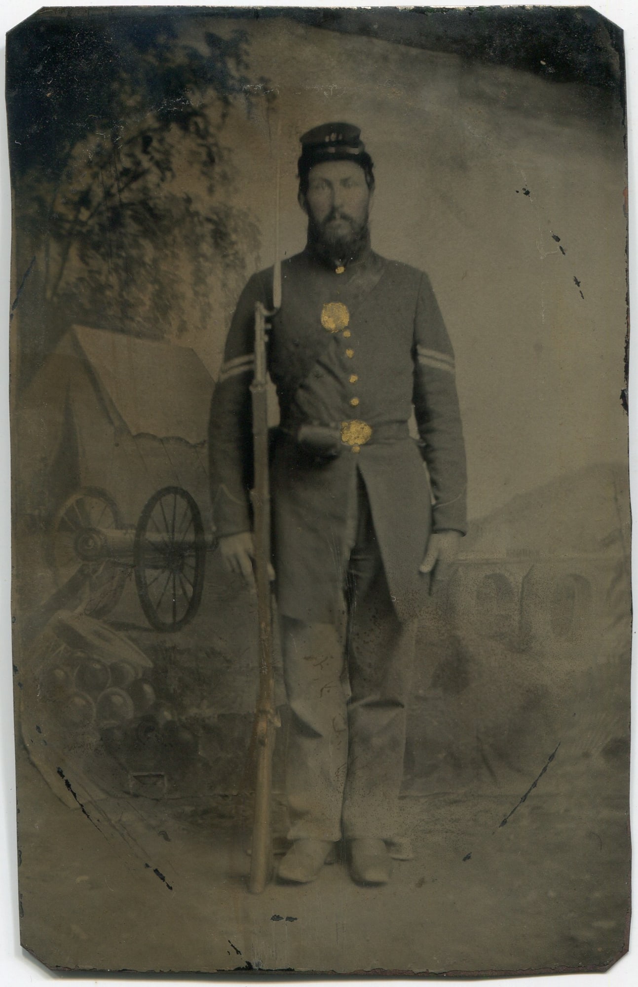 Tintype of Armed Civil War Soldier with Camp Backdrop: A quarter plate tintype depicting a Civil War soldier posed in full uniform with a rifle and fixed bayonet. The soldier stands with stoic confidence, wearing a kepi cap and dark coat adorned with hand