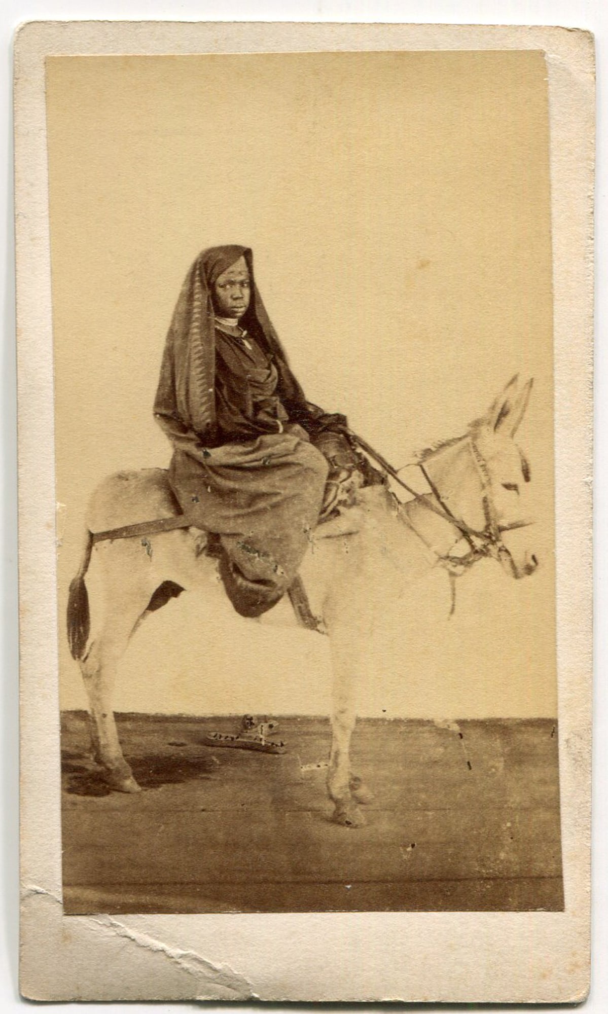 CDV of a Woman Riding a Donkey: An intriguing Carte de Visite (CDV) photograph captures a striking image of a woman riding a donkey. The subject is dressed in traditional attire, including a head covering that partially shields her 