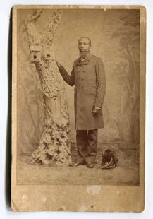 Cabinet Card of an African American Gentleman From Texas: Full-length cabinet card portrait of a well-dressed African American man posed beside a heavily textured faux tree trunk with carved-out birdhouses. His right hand rests on the tree while a soft cap o