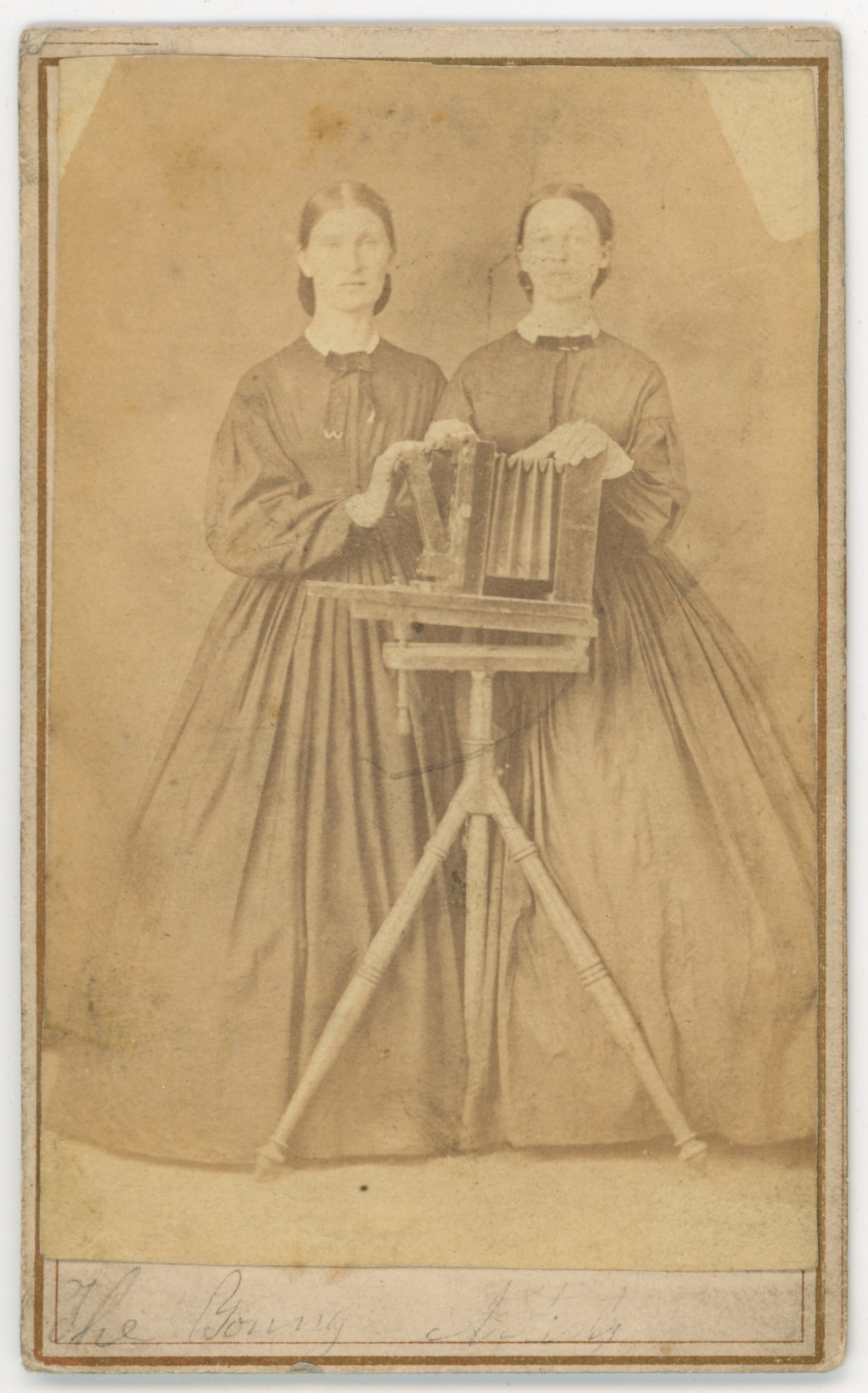 Two Women Photographers with Large Format Camera: Remarkable 1860s carte de visite depicting two women in matching dark dresses, posed in a studio with a large format bellows camera on a tall wooden tripod. Each woman holds part of the camera in a co