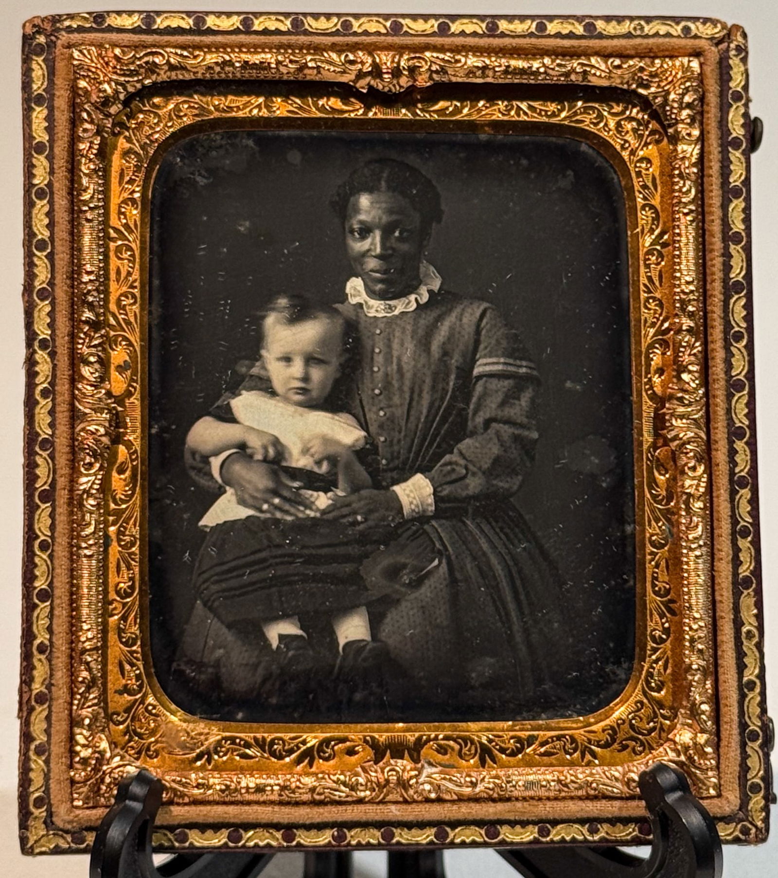 Daguerreotype of African American Woman with Identified Child: Sixth plate daguerreotype studio portrait depicting an African American woman seated and holding a white infant on her lap. The woman wears a patterned dress with a white ruffled collar, and the child