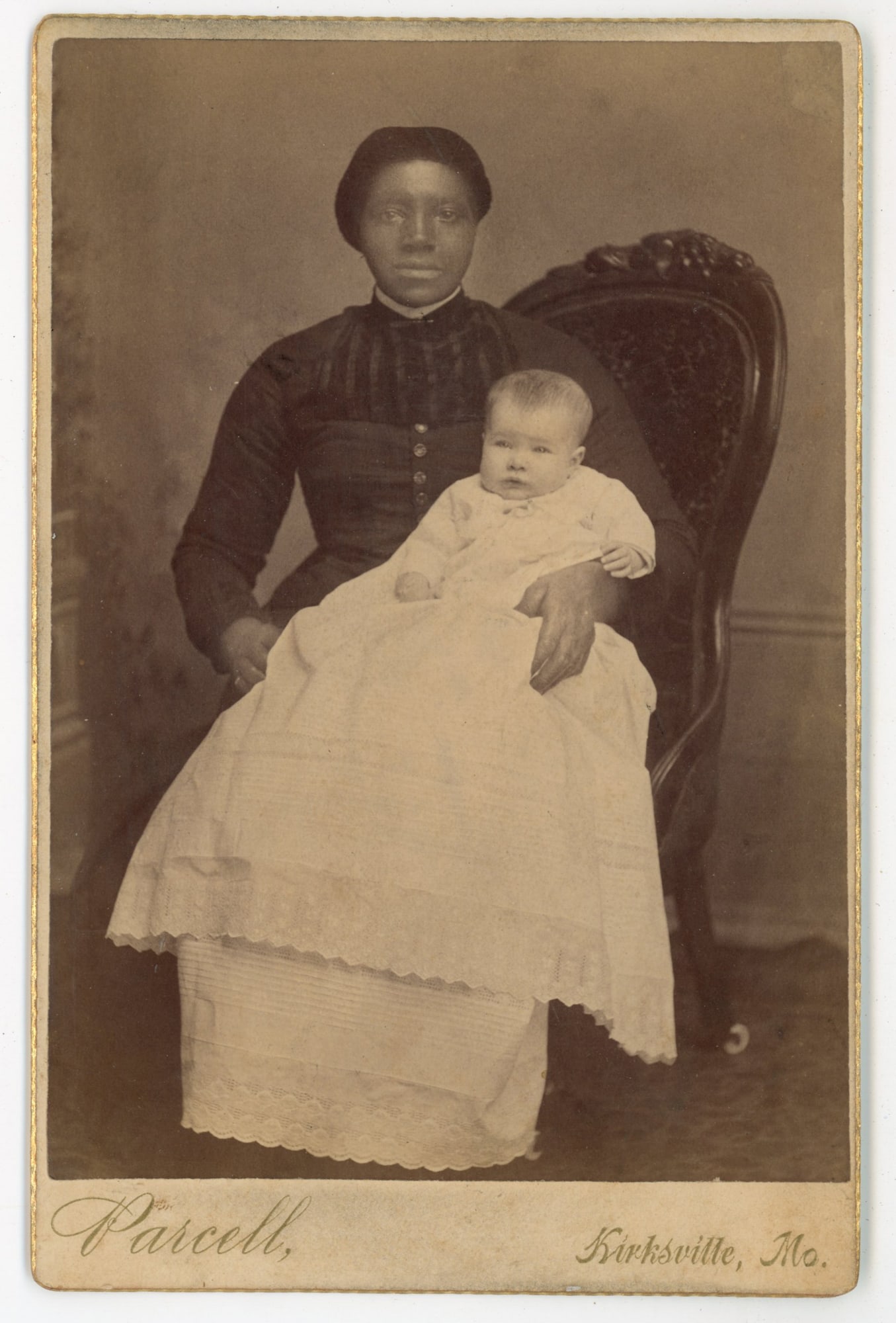 Cabinet Card African American Caregiver Holding White Infant, Kirksville, Missouri: Cabinet card by Parcell, Photographer, Kirksville, MO, ca. 1880s–1890s. A compelling studio portrait of a young African American woman seated in a carved Victorian chair with a white infant in a lon