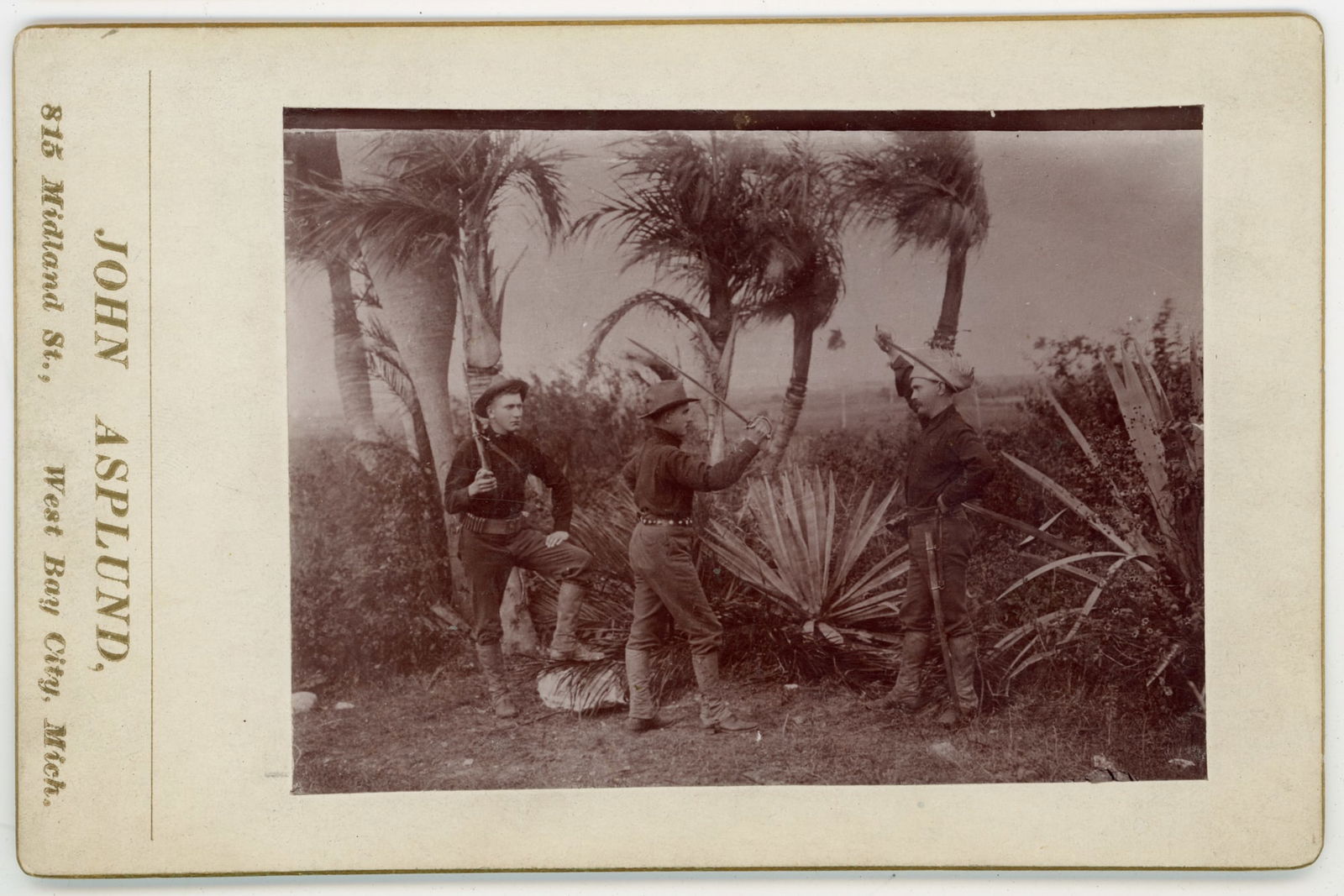 Spanish–American War Soldiers in Tropical Camp, Albumen Print: Albumen print depicting three U.S. soldiers posed with rifles in a tropical outdoor setting, likely during the Spanish–American War period, circa 1898. The men stand among palm trees and broad-leafe