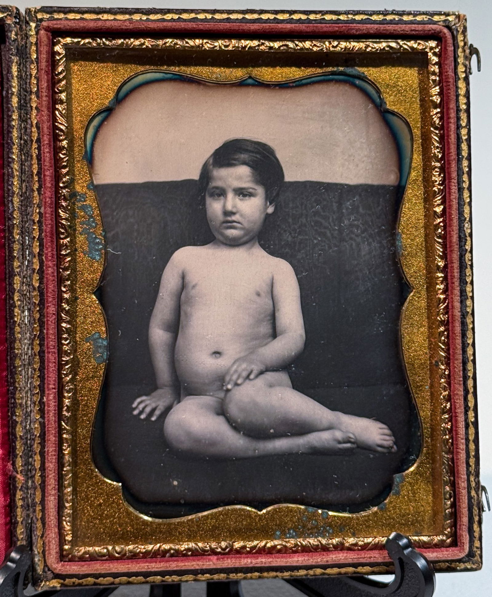 Daguerreotype of Nude Child Seated on Cushion: Sixth plate daguerreotype studio portrait depicting a nude young child seated on a dark upholstered cushion, posed in a relaxed yet deliberate position with one leg extended and the other bent beneath