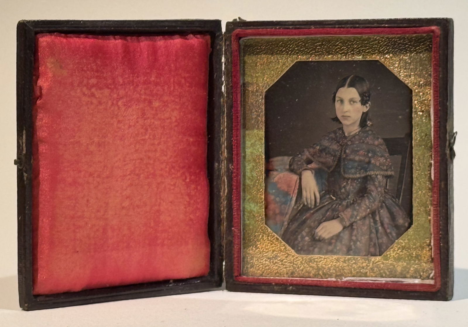 Daguerreotype Portrait of Young Woman with Elaborately Tinted Dress: Sixth plate daguerreotype studio portrait of a young woman seated beside a table, posed in three-quarter view and looking directly toward the camera. The image is a daguerreotype on a silvered copper
