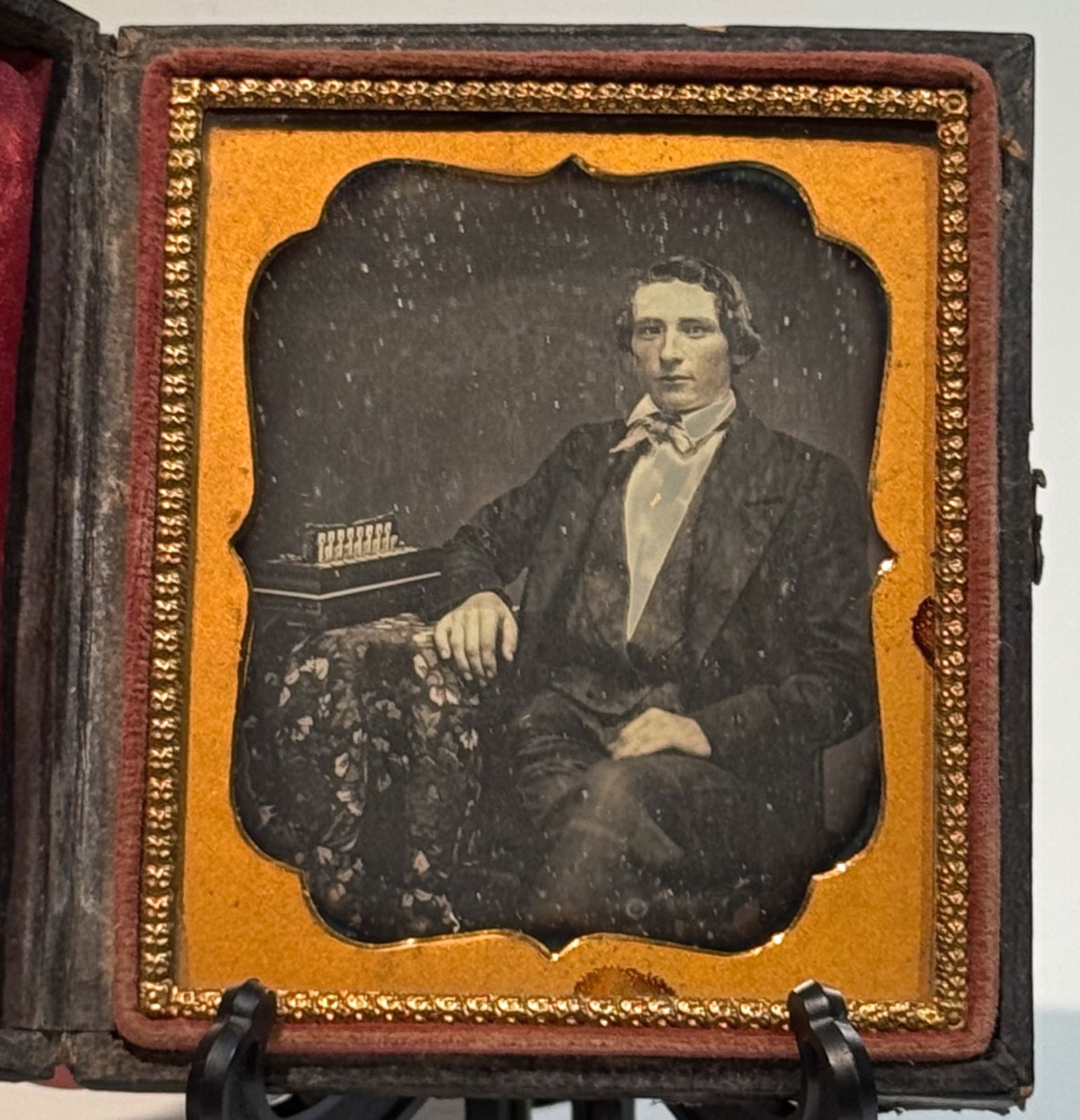 Daguerreotype Portrait of Man with Concertina: Sixth plate daguerreotype studio portrait of an unidentified man seated beside a small table, holding a concertina. The subject wears a dark jacket with a light shirt and cravat, and is posed in a for