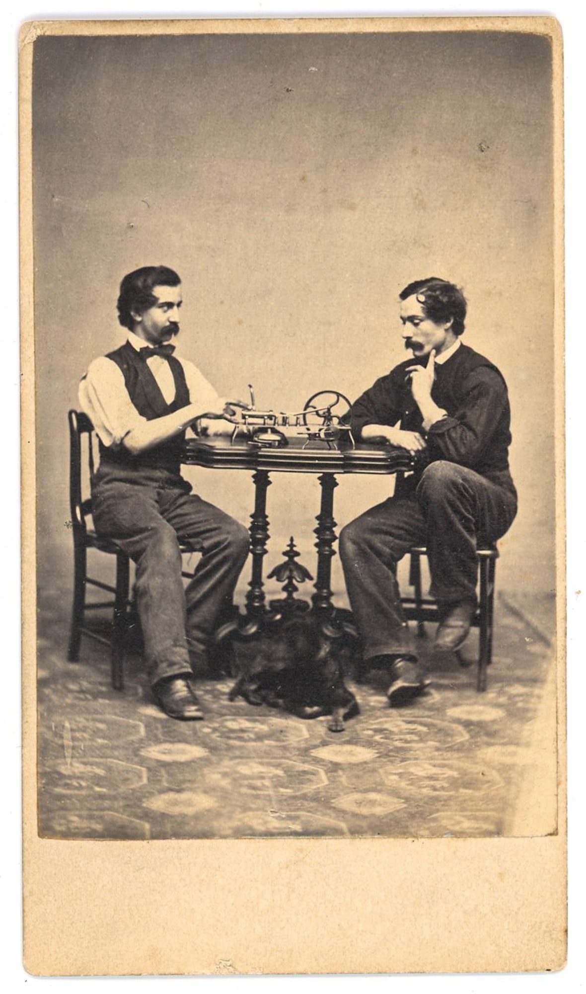 CDV Studio Scene with Telegraph Apparatus, Two Men at Table: Carte-de-visite photograph depicting two adult men seated at a small table with a telegraph apparatus prominently placed between them. One man operates a telegraph key while the other observes in a co