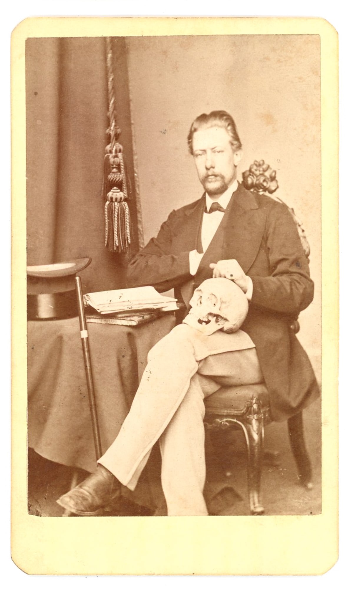 CDV Portrait, Unidentified Man with Skull Prop: Albumen carte-de-visite studio portrait of an unidentified adult man seated in formal dress, posed with a human skull resting across his lap. The theatrical studio setting includes a draped table with