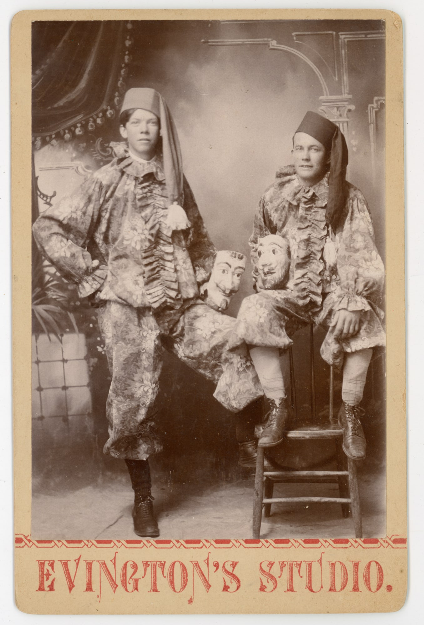 Masqueraders with Masks, Cabinet Card Albumen Print: Cabinet card albumen print depicting two young men posed in elaborate masquerade costumes, each wearing patterned theatrical attire and soft caps, with carved masks incorporated into the composition.