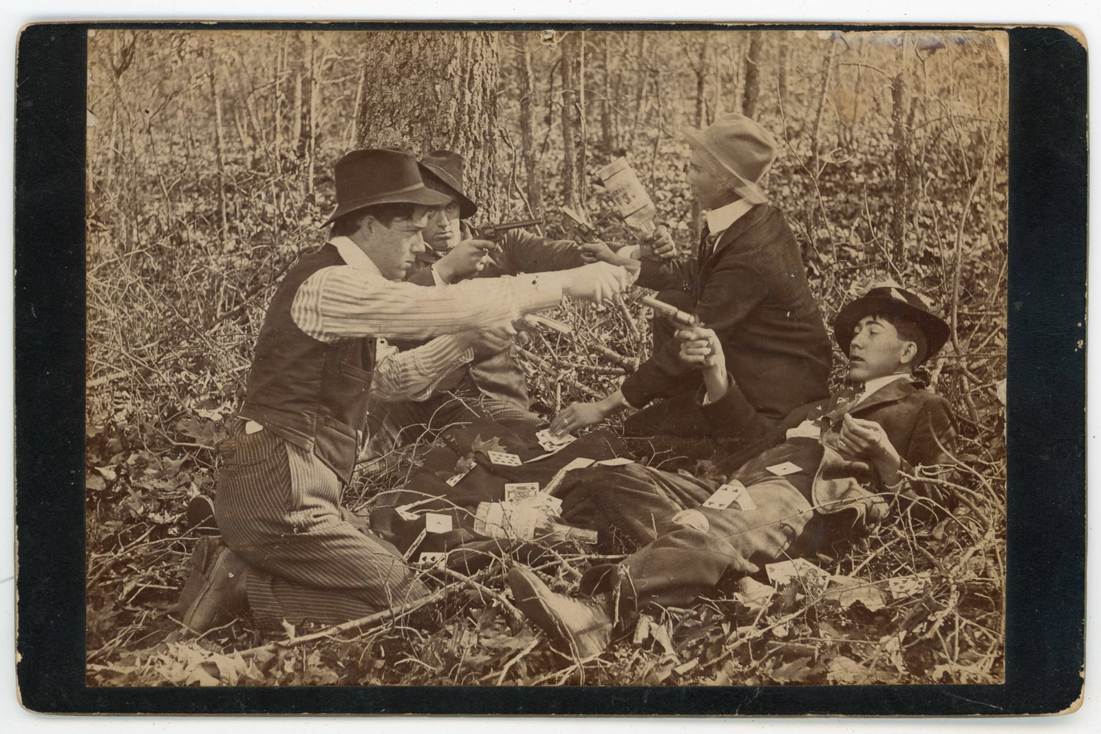 Cabinet Card Poker Scene Shoot Out: Albumen cabinet card depicting a staged gambling scene in which a casual card game escalates into a gunfight. Four young men are shown outdoors in a wooded setting, seated and reclining on the ground