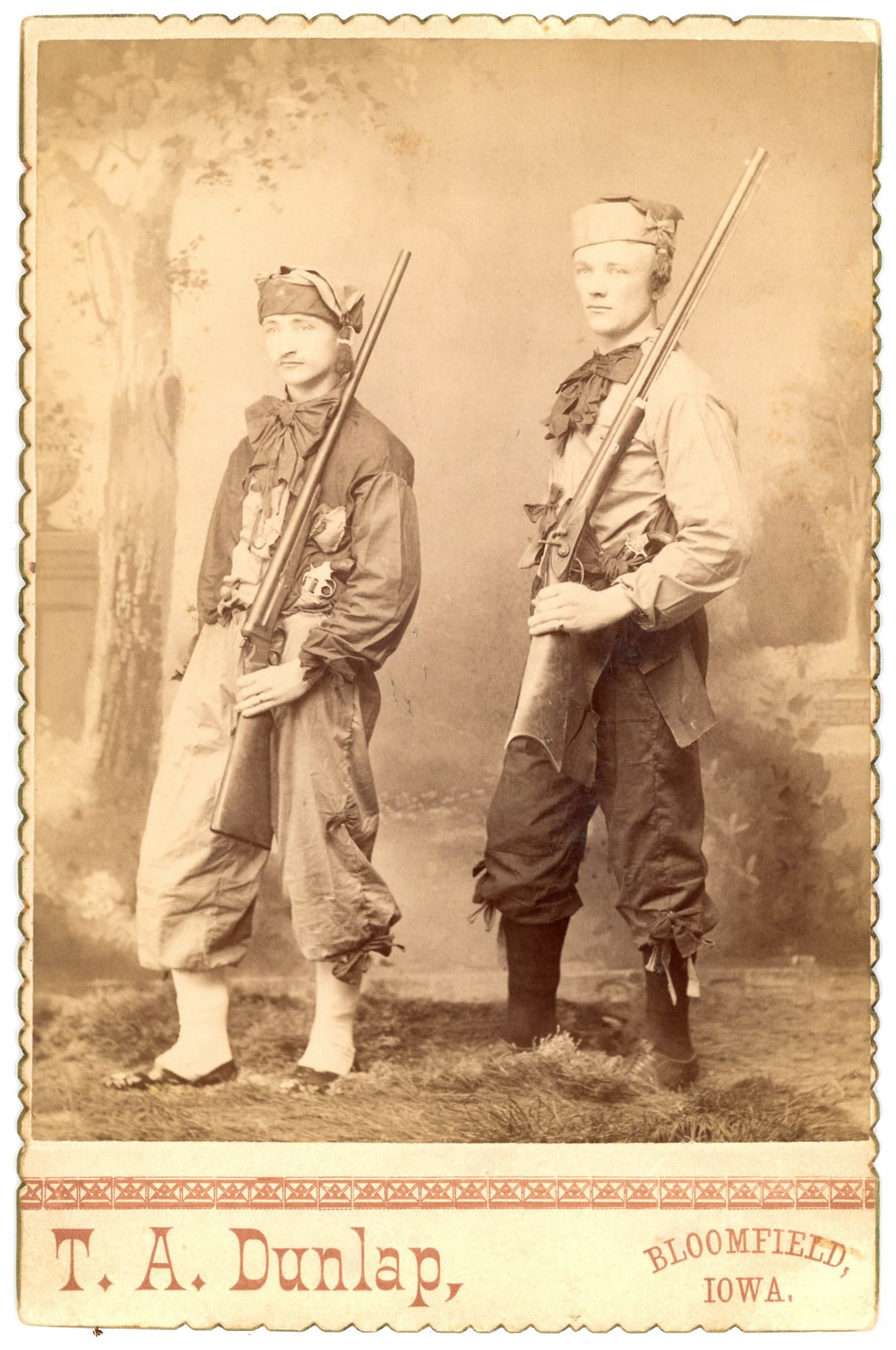 Cabinet Card, Two Men with Rifles, Bloomfield, Iowa: Cabinet card studio portrait depicting two adult men posed standing side by side, each holding a long rifle. The men wear matching or coordinated frontier-style clothing, including loose trousers tied