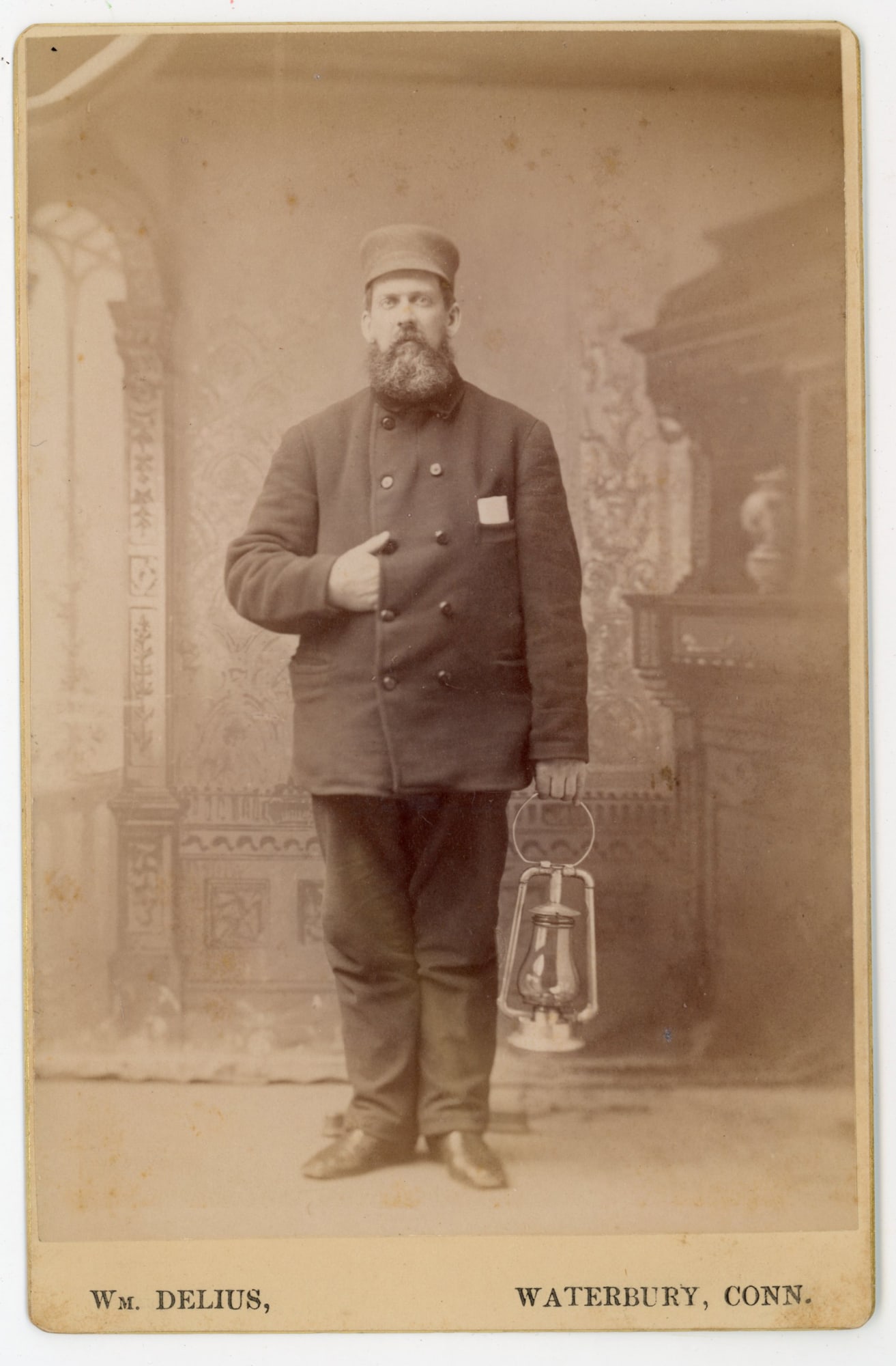Railroad worker with Lantern Portrait Waterbury CT 1887: Cabinet card photograph depicting a long-haired Skye Terrier seated inside a woven wicker basket with the lid propped open behind the dog. The small terrier’s long coat falls over its eyes and down
