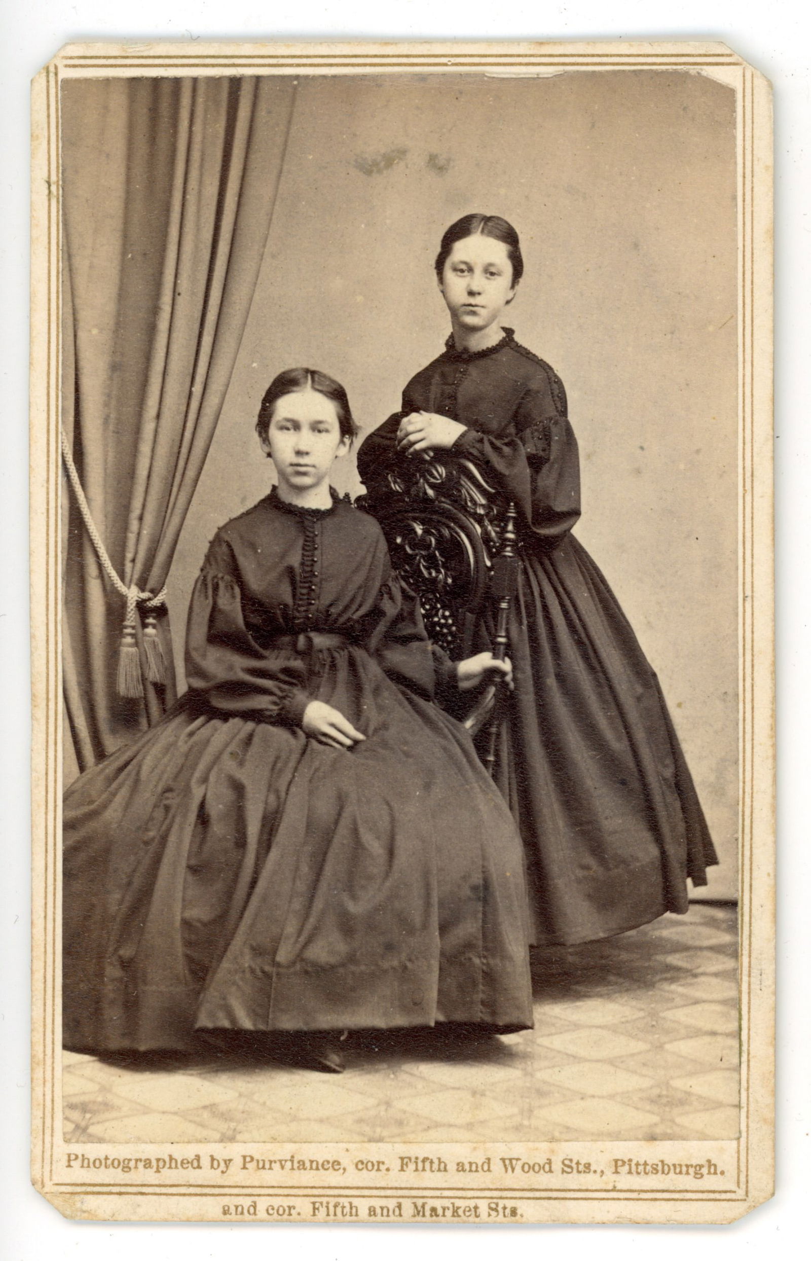 CDV Women Twins Portrait Purviance Pittsburgh: Carte de visite studio portrait depicting twins posed together in formal mid-nineteenth century dress. One woman is seated while the other stands beside an ornate studio chair within a curtained inter