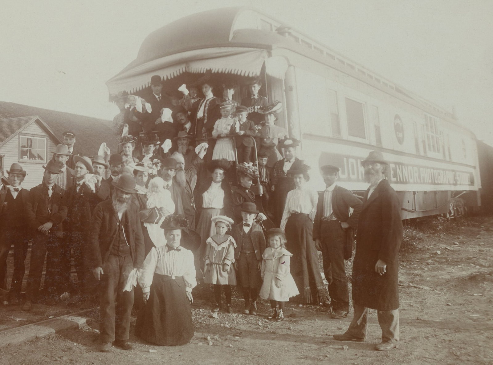 Mounted Photograph John Ennor Photographic Railcar: Albumen photograph showing a large group posed at the rear platform of a railroad photographic studio car marked John Ennor Photographic Studio, circa 1890s–1900s. The image shows men, women, and ch