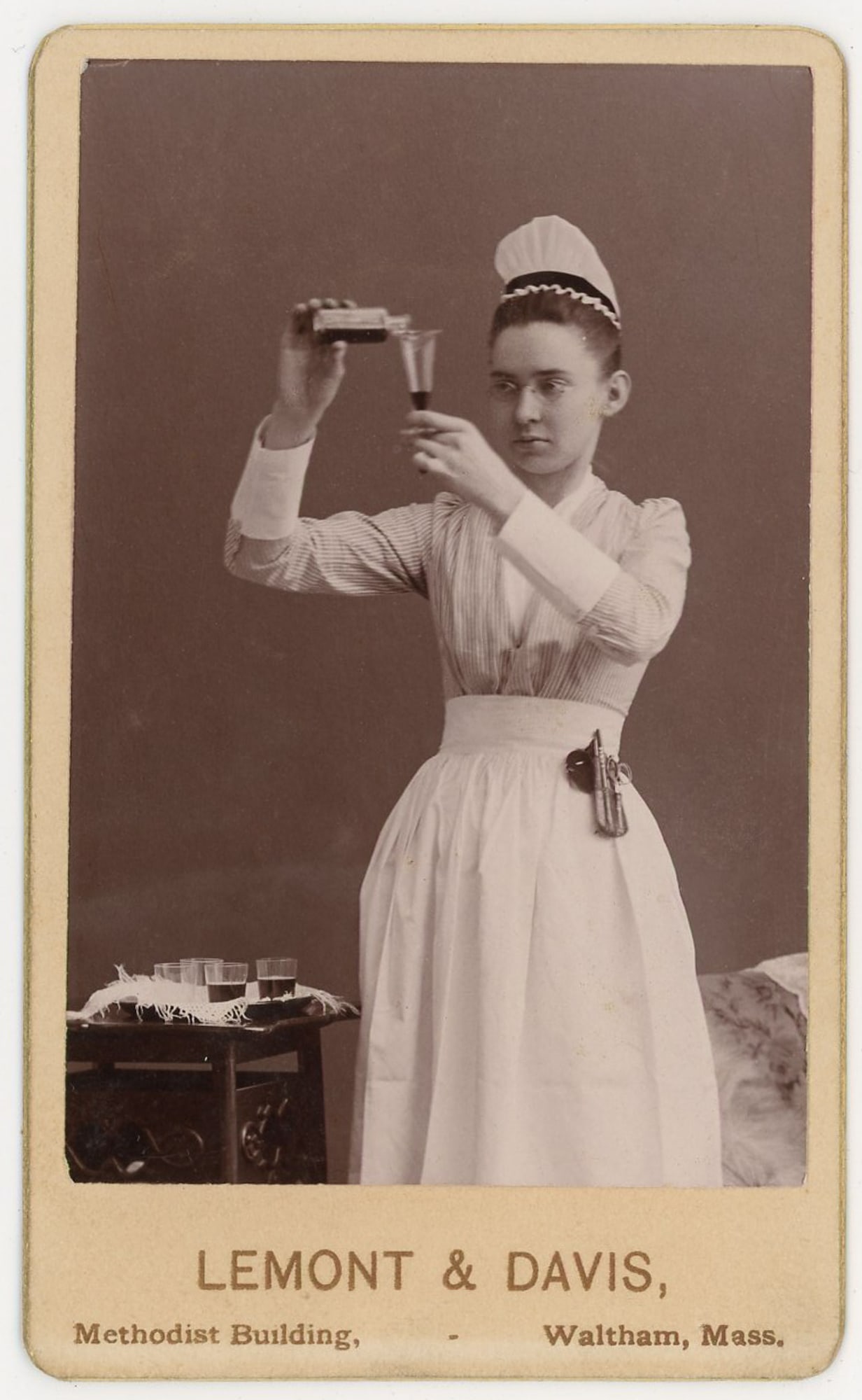 CDV Nurse Measuring Medicine, Waltham MA: CDV photograph depicting a young nurse or medical assistant holding a vial and carefully pouring a dark liquid into a small glass for measurement. The subject wears a nurse’s cap and uniform and is