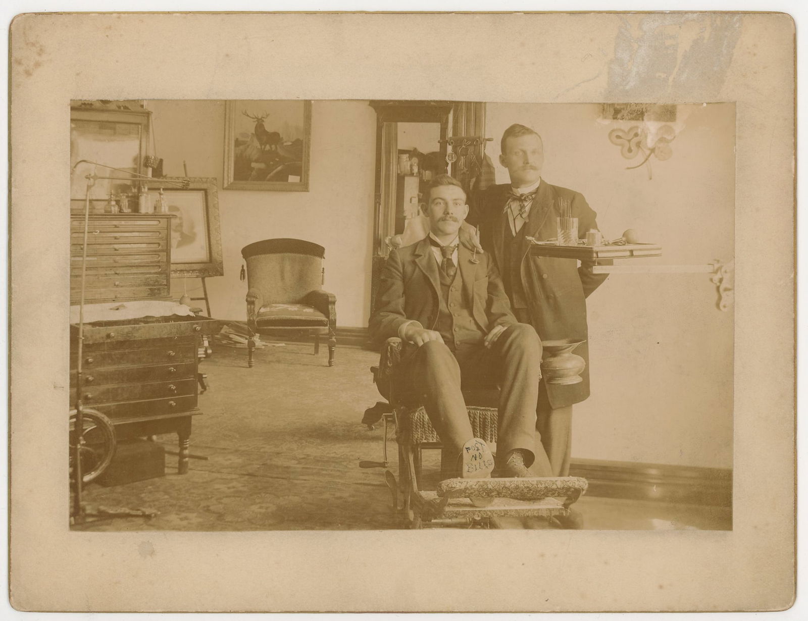 Gelatin Silver Photograph Dental Office Interior: Interior photograph showing a dental office with two men posed among equipment and furnishings, gelatin silver print dating to the early 20th century. One man is seated in a dental chair while the oth