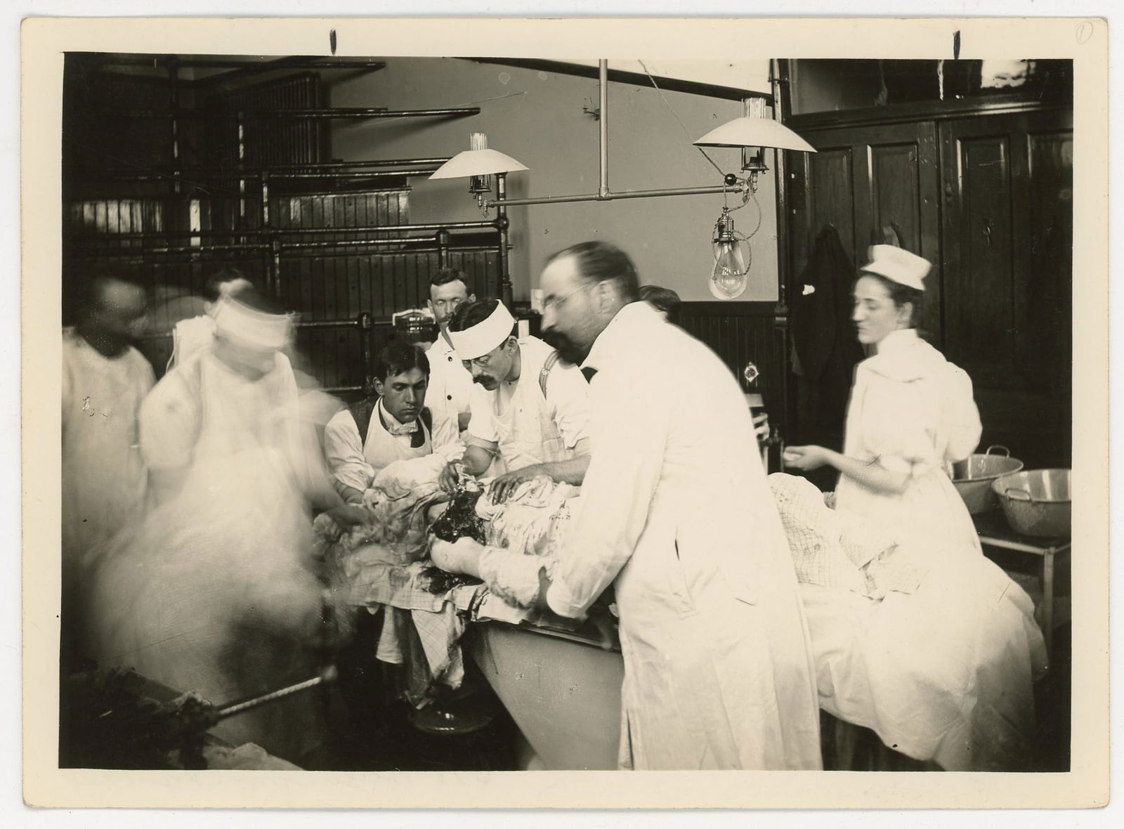 Operating Room Surgery Photograph, Early Medical Scene: Silver gelatin photograph showing a surgical team performing an operation in a hospital operating room. Several doctors and assistants in white coats and caps surround the patient on the operating tab