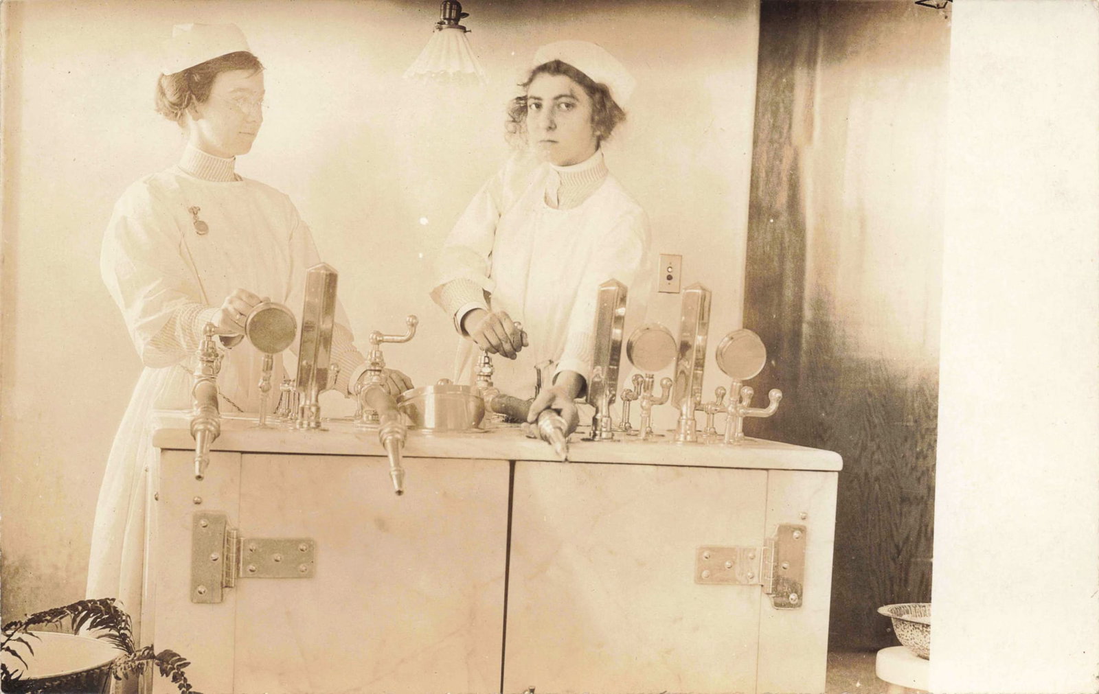 RPPC Nurses with Anestheology Equipment: Real photo postcard showing two nurses posed beside a large cabinet with multiple taps and gauges apparently related to early use of anestheology. Real photo postcard stock. No mailed use is visible;