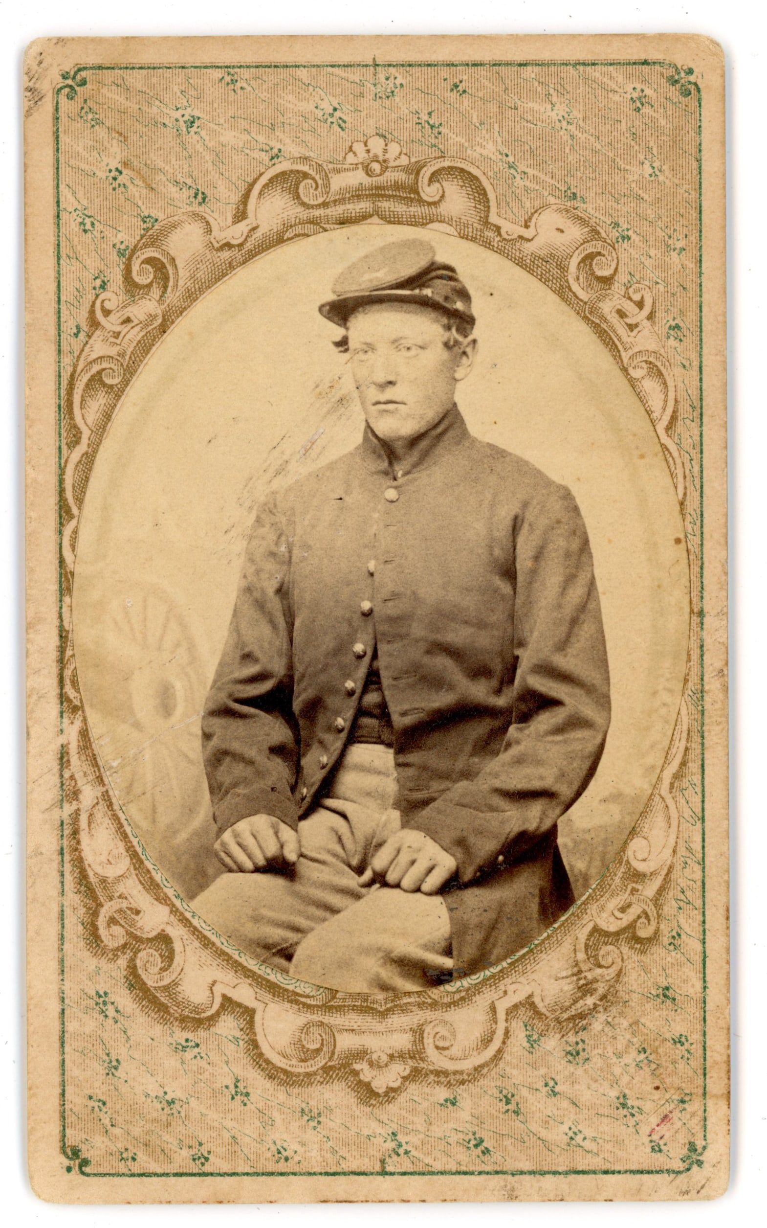 CDV Civil War Soldier Oval Portrait: Carte de visite portrait depicting an unidentified Union Civil War soldier in uniform presented within an oval vignette on a decorative printed mount. The soldier is shown seated wearing a military co
